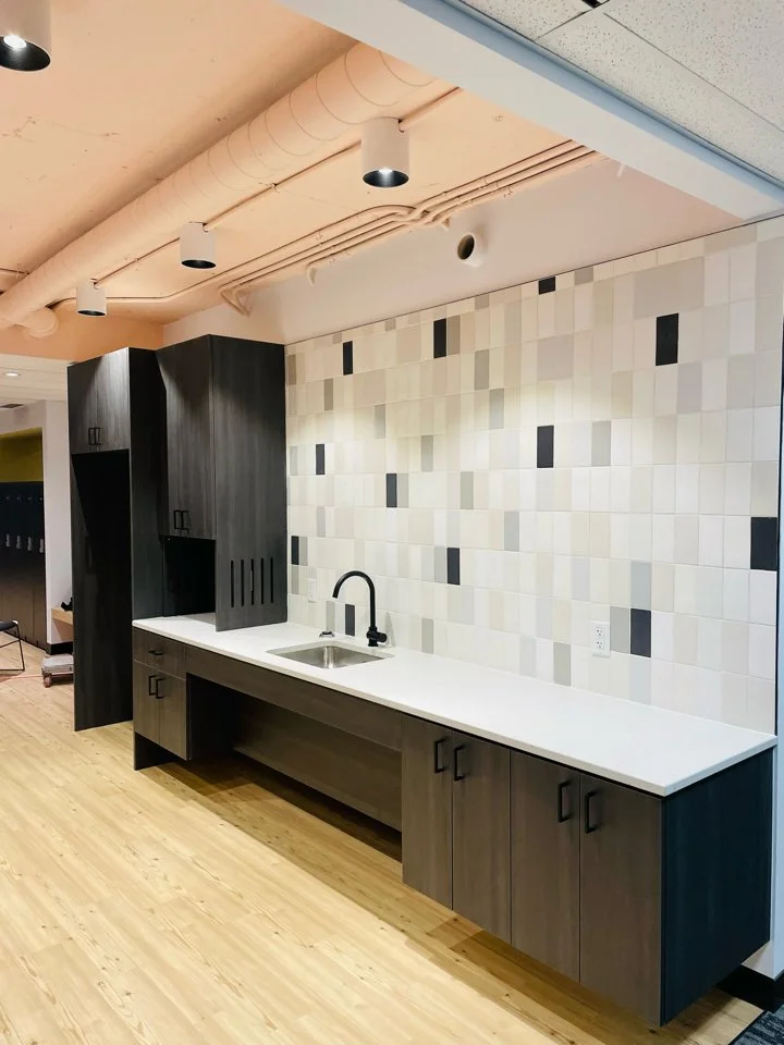 Modern kitchenette with dark wood cabinets, a white countertop, and a beige patterned tile backsplash in an interior space.