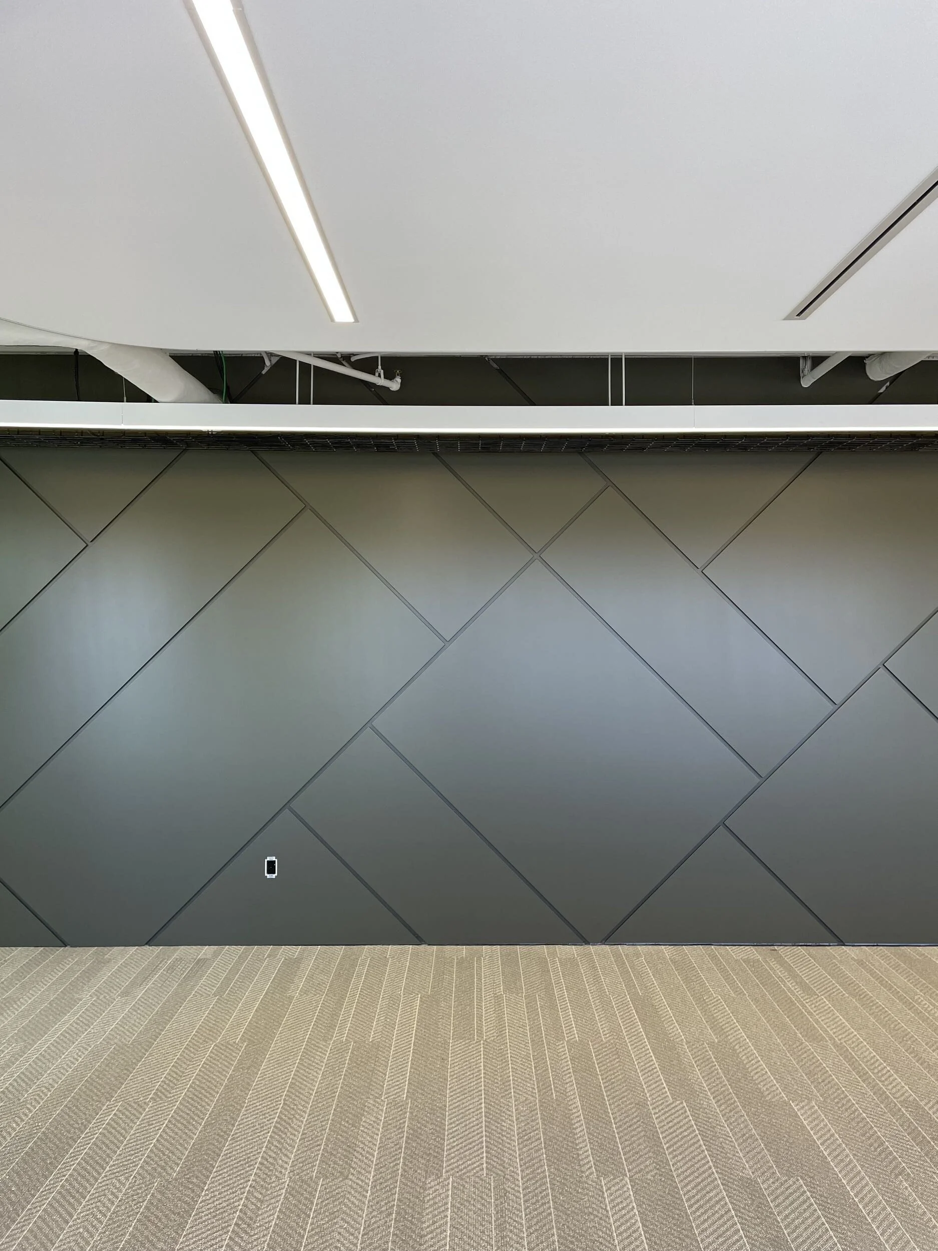 Interior view of an office or airport with a large gray wall, a grid of tiles on the floor, and a ceiling with white panels and fluorescent lights.