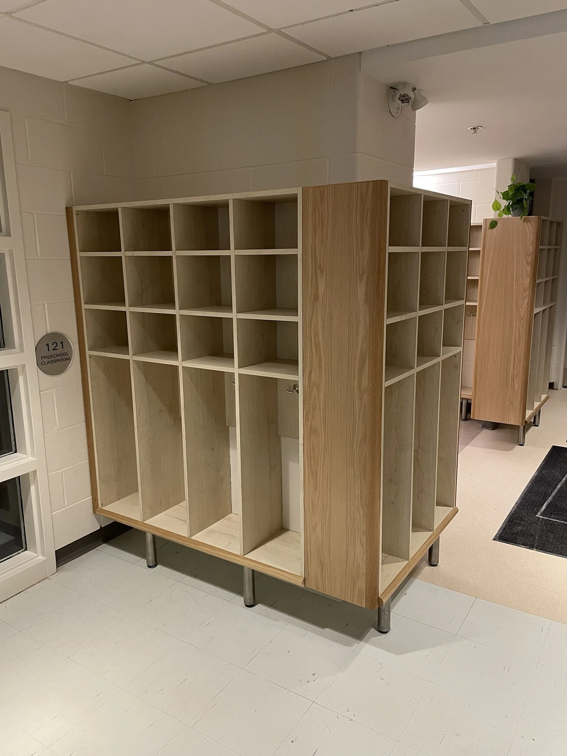 Empty wooden cubby shelving unit in corridor, with small plant on top, in a school or daycare environment.