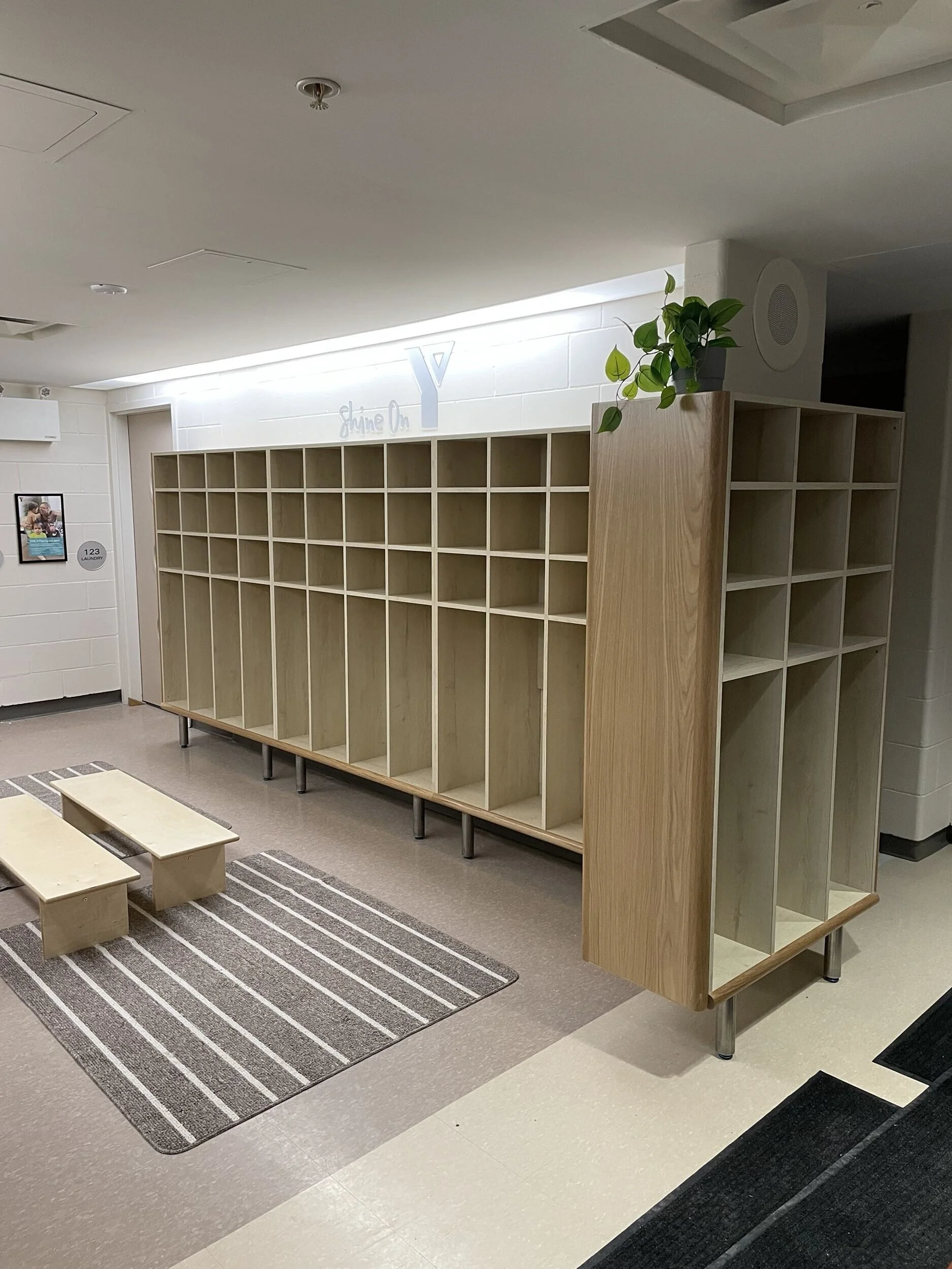 Empty wooden cubby storage unit in an indoor public space with a small plant on top and a sign that says 'Shine On Y' in the background.