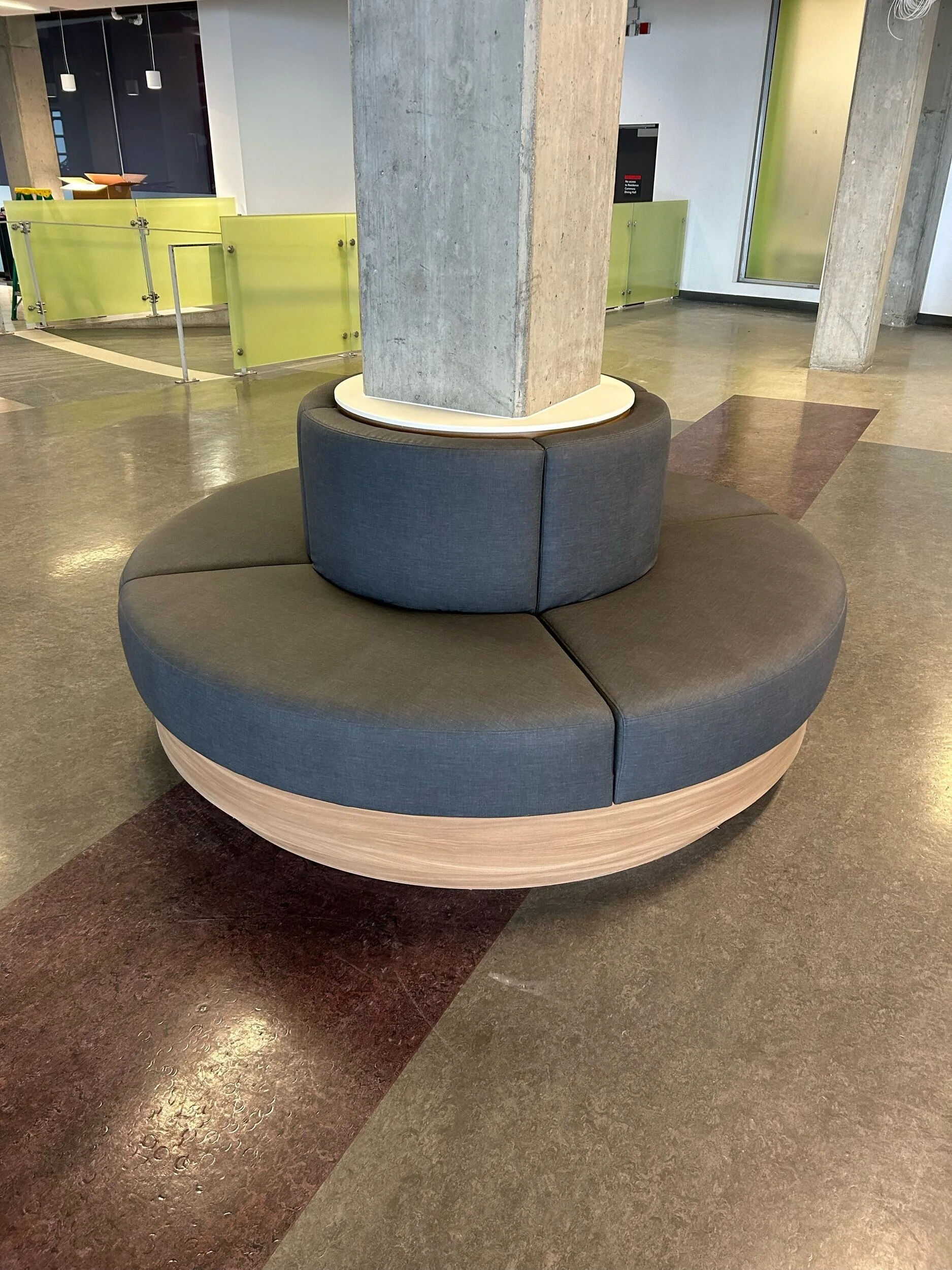 A circular gray seating area with a tall, rectangular concrete pillar in the center, situated in a modern interior space with colorful flooring and green wall accents.
