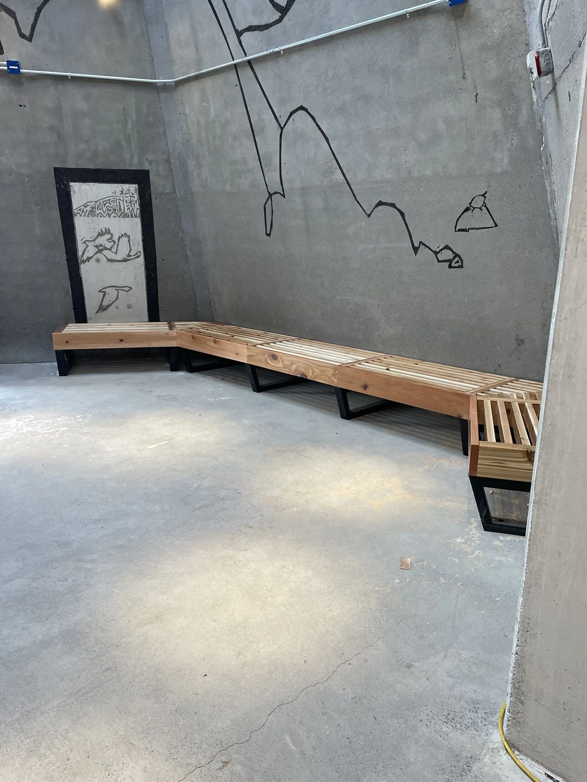 Interior view of a concrete room with a long wooden bench along the wall, a hanging abstract mural of a bird on a white door, and a black line drawing of mountain peaks on the wall.