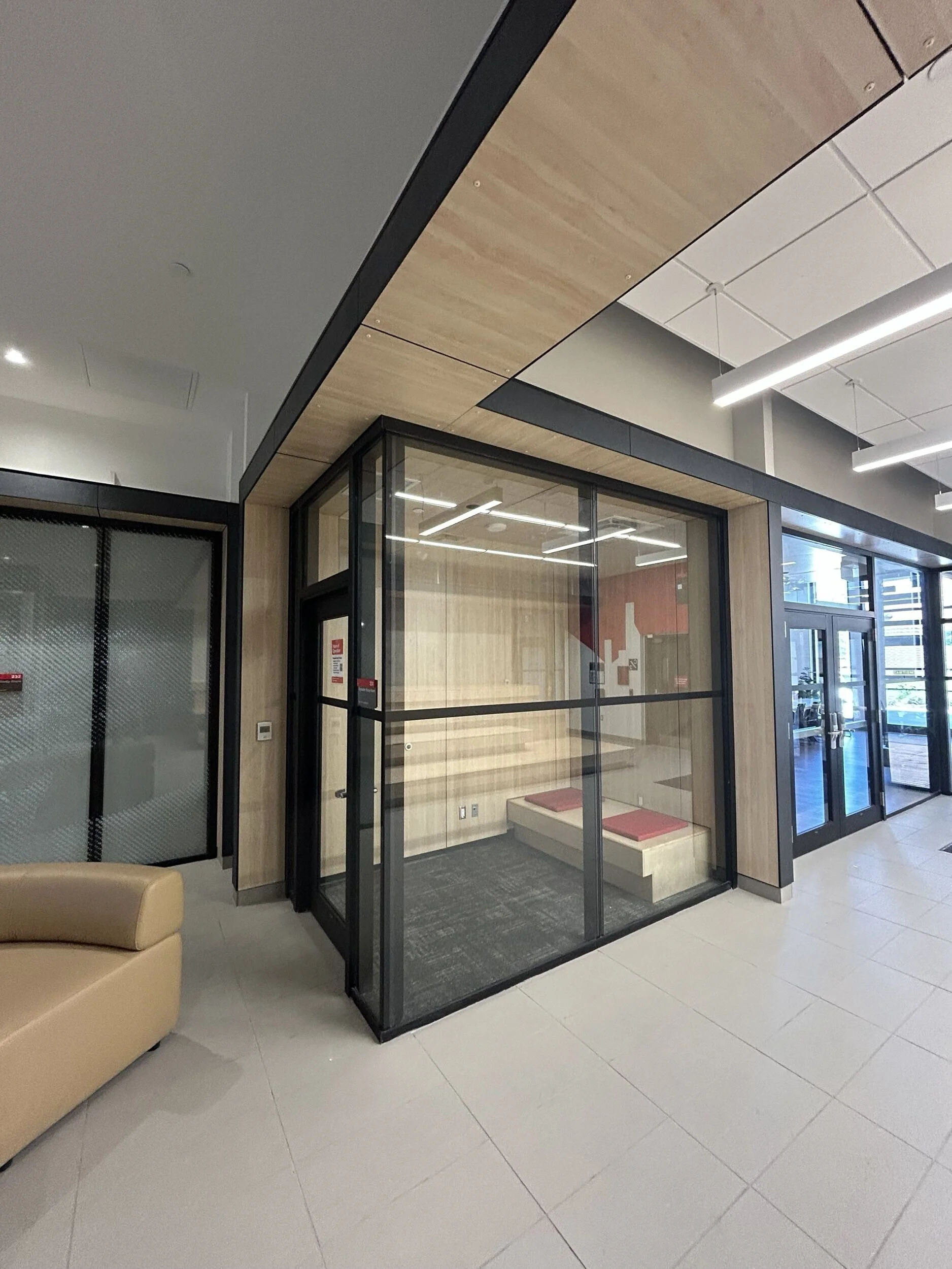 Inside a modern building with a glass-enclosed room, a beige couch, and tiled flooring. There are ceiling lights and a child-sized bench inside the glass room.