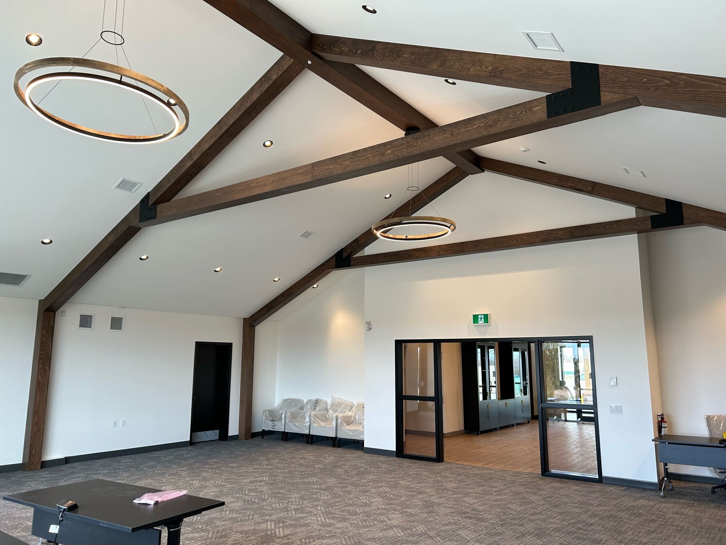 Interior view of a spacious room with modern ceiling design featuring exposed wooden beams and circular ring pendant lights. The room has white walls, recessed lighting, and a carpeted floor. There are chairs covered in plastic, a black table, and la