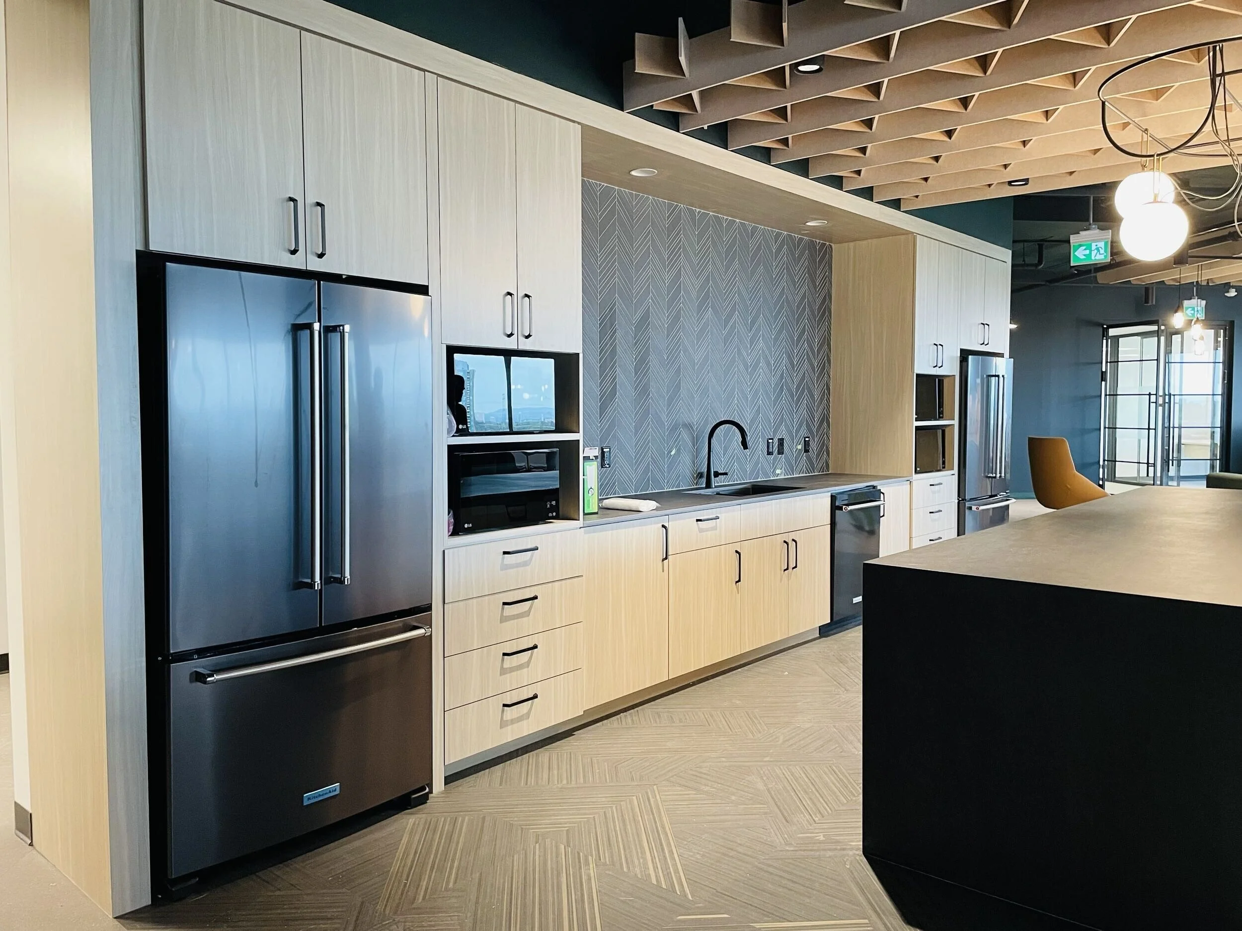 Modern kitchen with light wood cabinetry, stainless steel refrigerator, microwave, and black sink. Textured blue feature wall, wood ceiling panels, and pendant lights in the background.