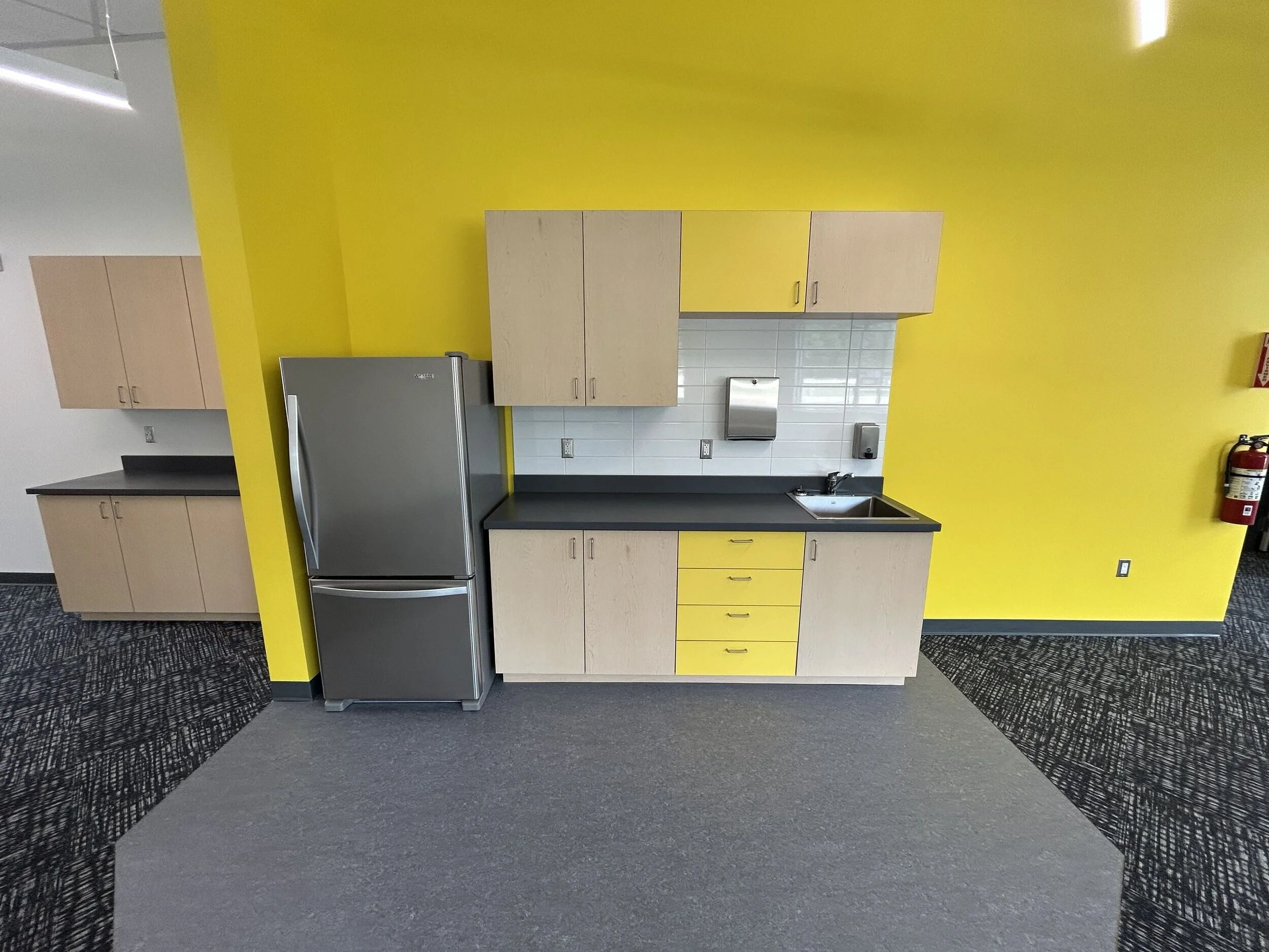 A bright yellow wall in a modern kitchen with light wood cabinets, a stainless steel refrigerator, a white tile backsplash, a sink, and a fire extinguisher mounted on the wall.