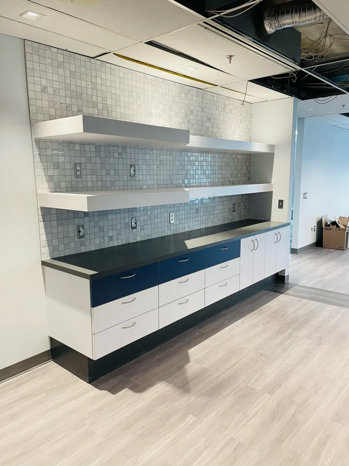 Empty kitchen or break area with white and blue cabinets, three open white shelves, gray tiled backsplash, and electrical outlets.