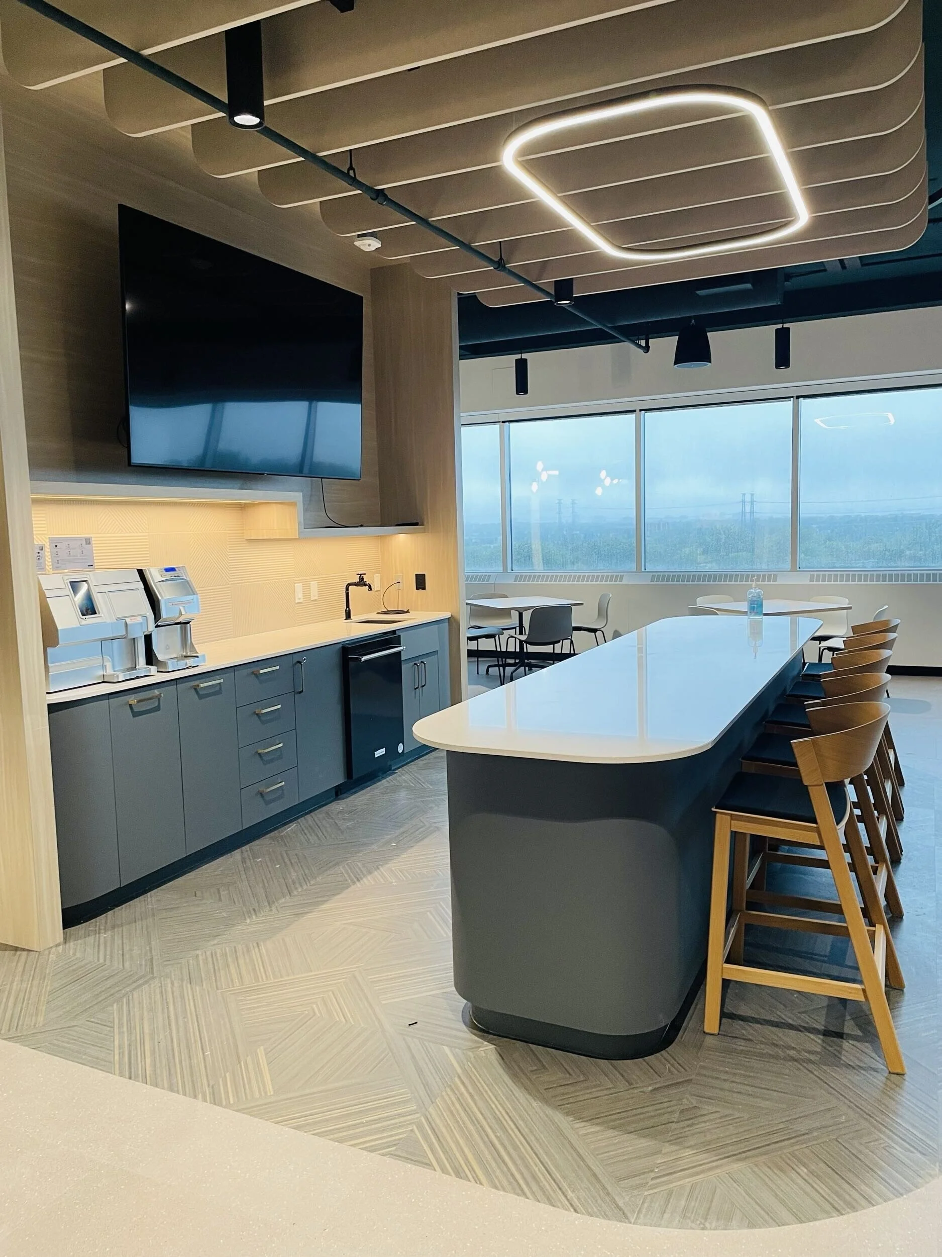 Modern lounge or kitchen area with a large white island counter, wooden bar stools, a blue cabinet with coffee machines, a large wall-mounted TV, and a window showing an overcast sky and power lines outside.