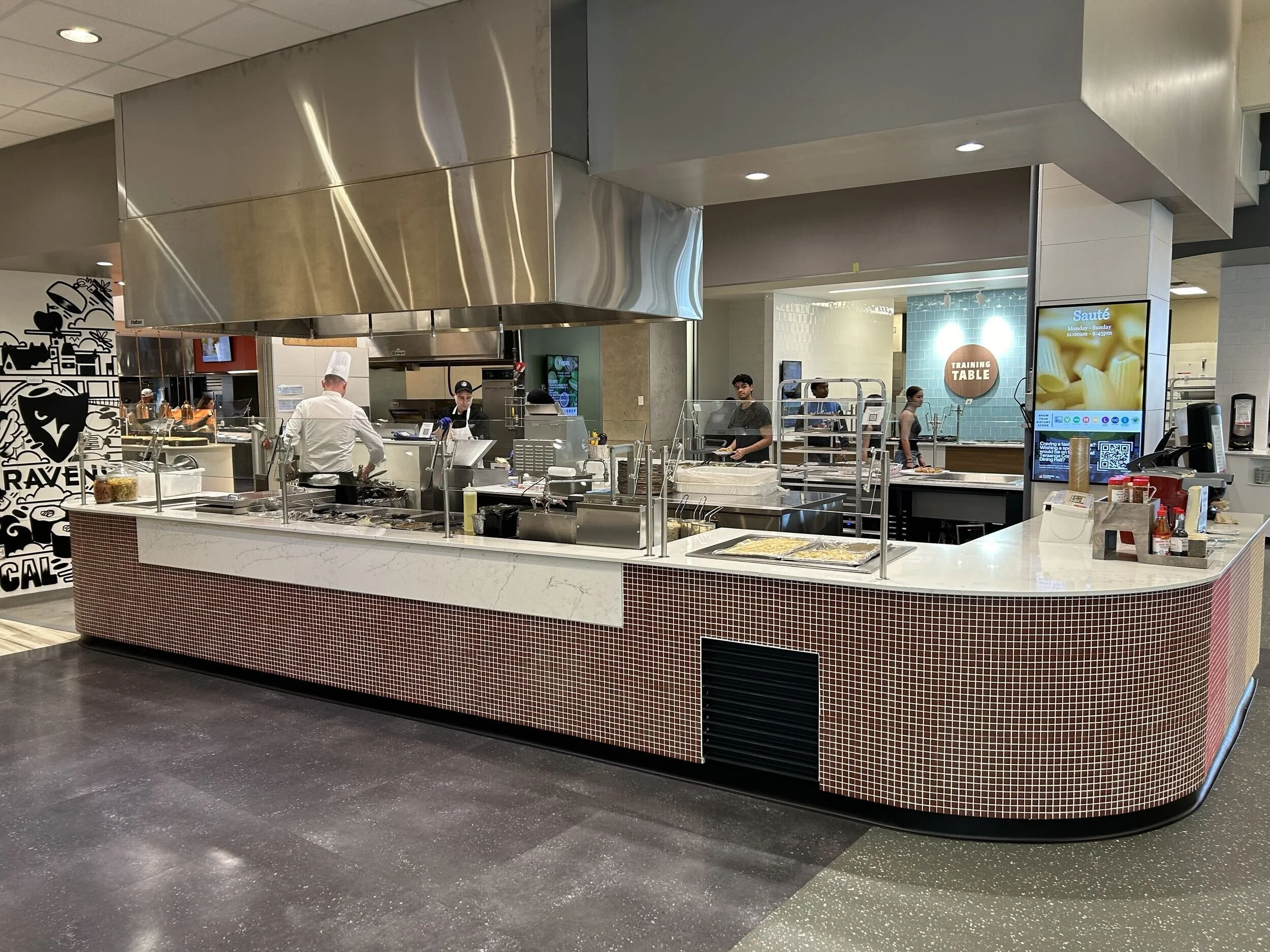 A food court counter with a stainless steel hood, kitchen area, and several staff members working, including chefs and servers. Digital menu boards and a digital screen display menu options and promotional content.