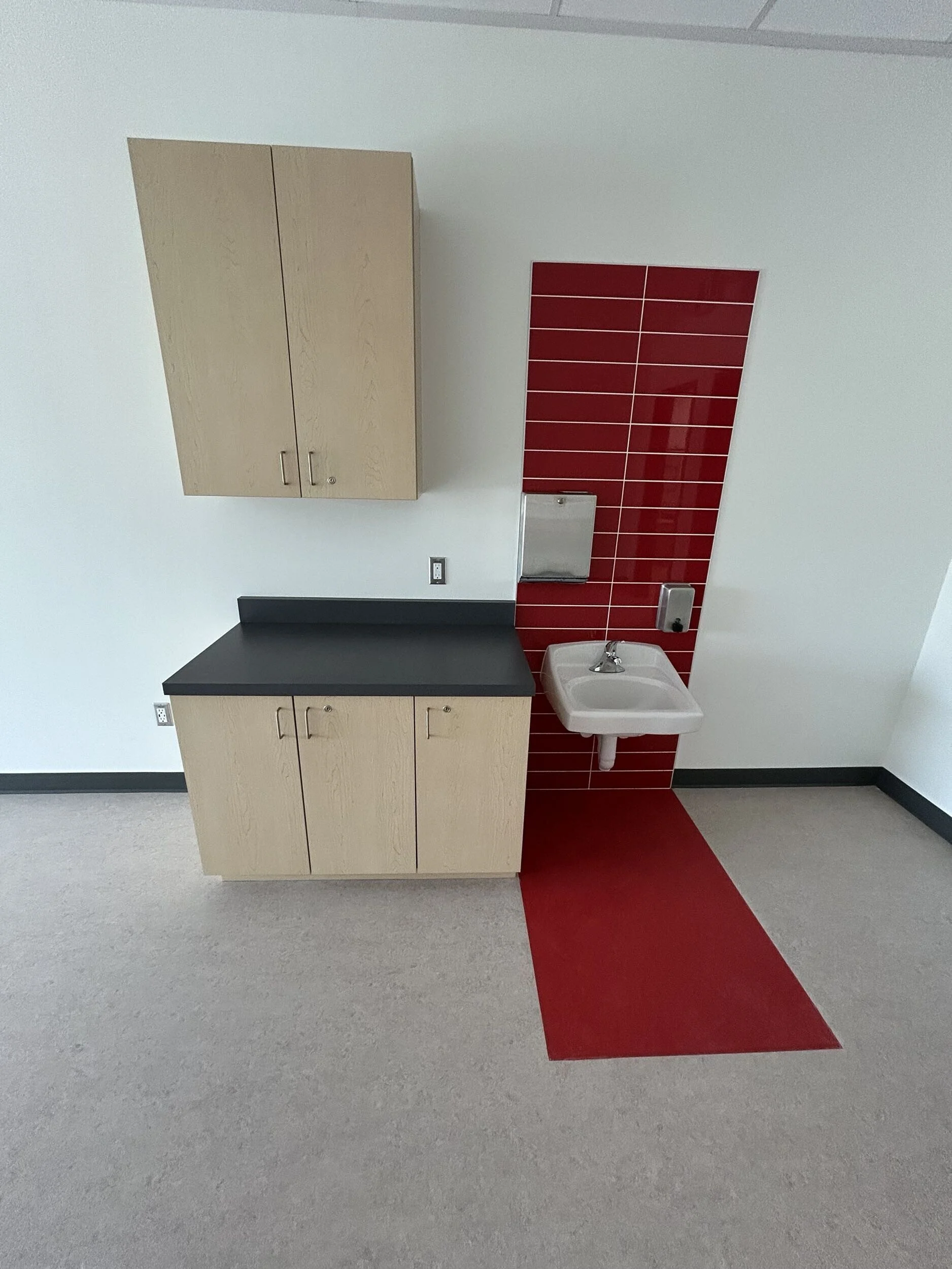 A corner of a room with a red tiled wall, a white sink, a wall-mounted paper towel dispenser, a wooden cabinet, and a gray and red floor.
