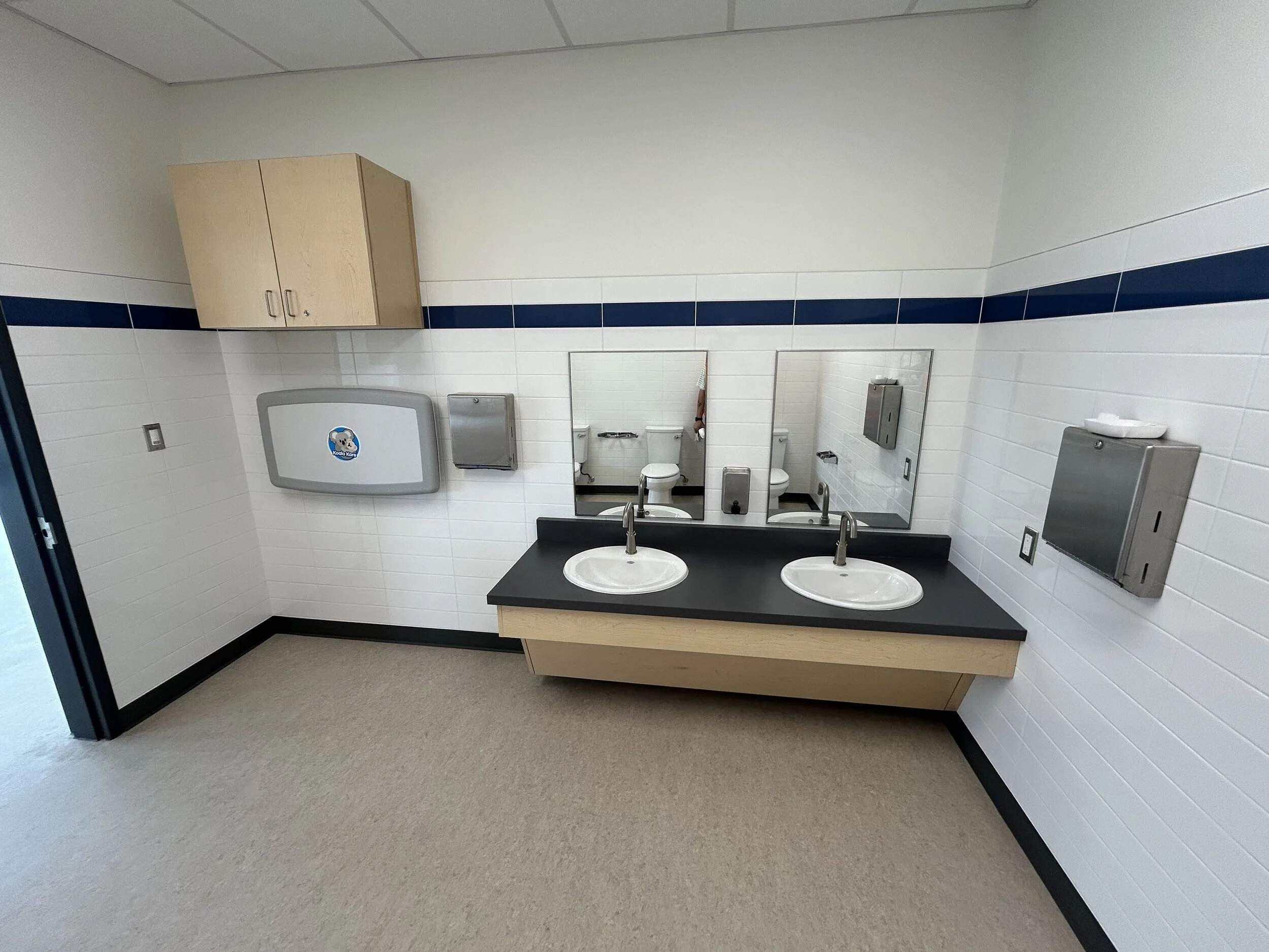 Public restroom with double sinks, mirrors, paper towel dispenser, soap dispenser, hand dryer, and bathroom stalls with closed doors.