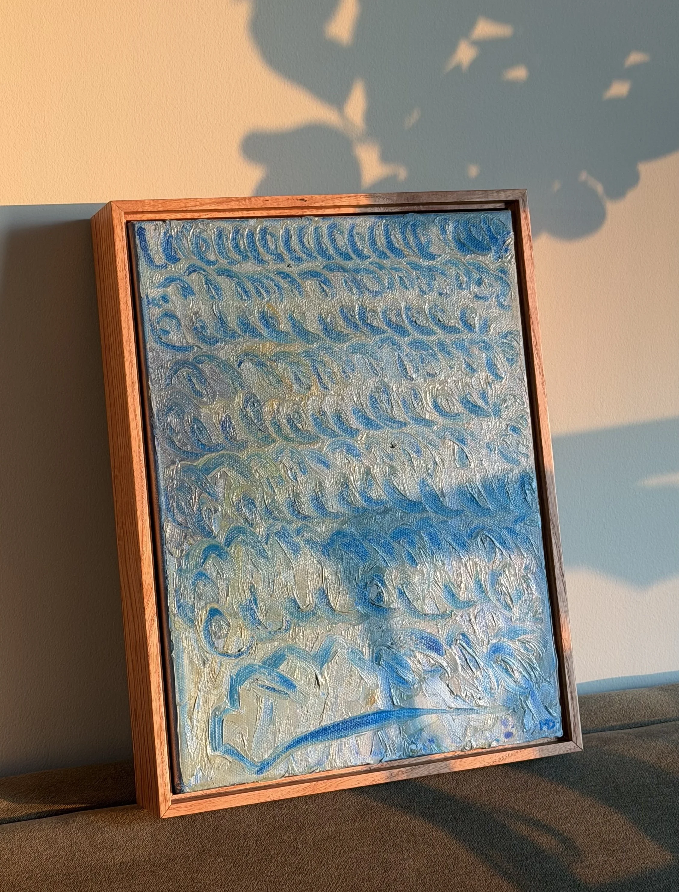 Framed abstract painting with swirling blue and yellow brushstrokes, leaning against a beige wall, illuminated by natural light, casting a shadow on the wall.