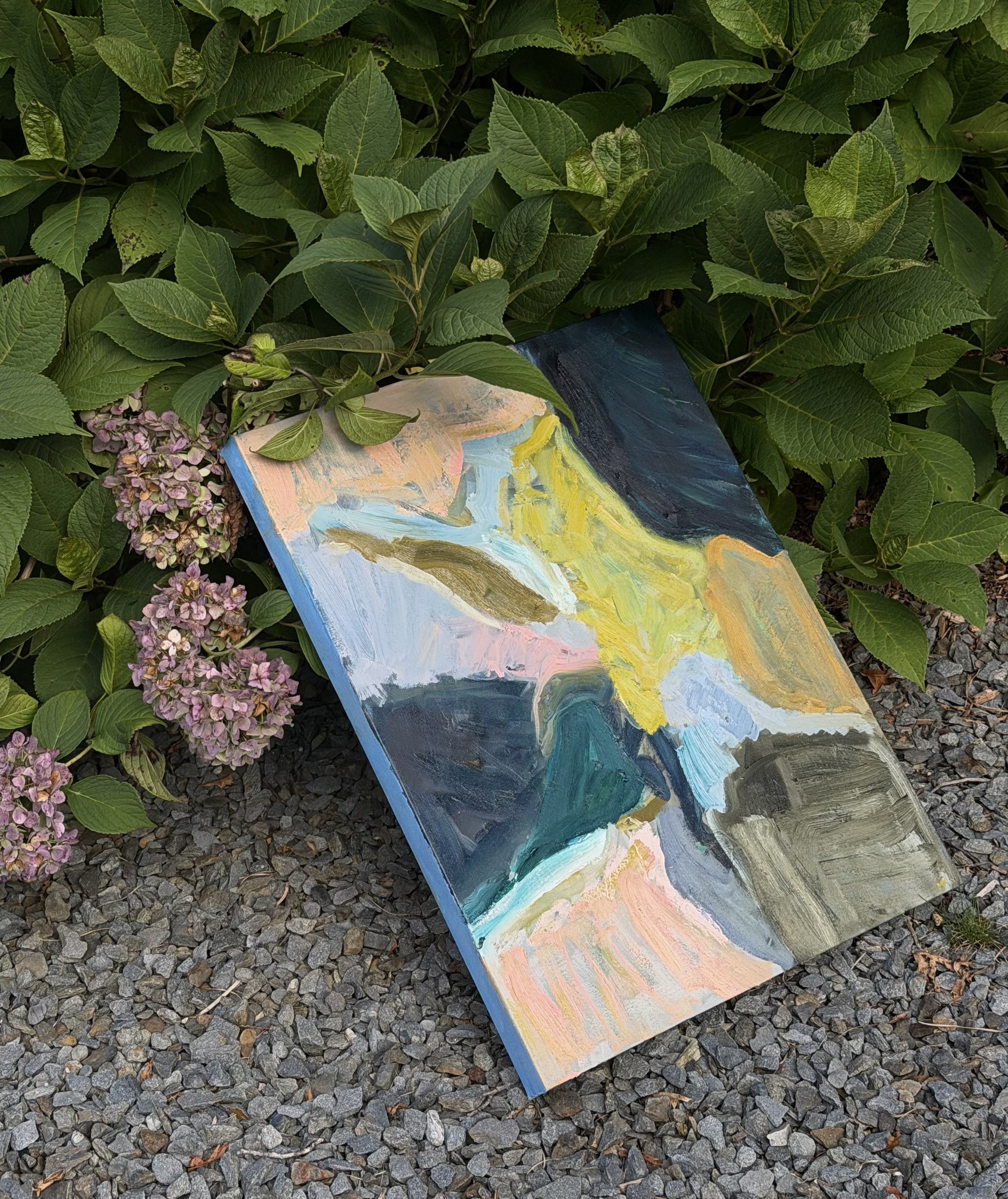 A colorful painted canvas lies on the ground amidst green leafy bushes and pink hydrangea flowers.