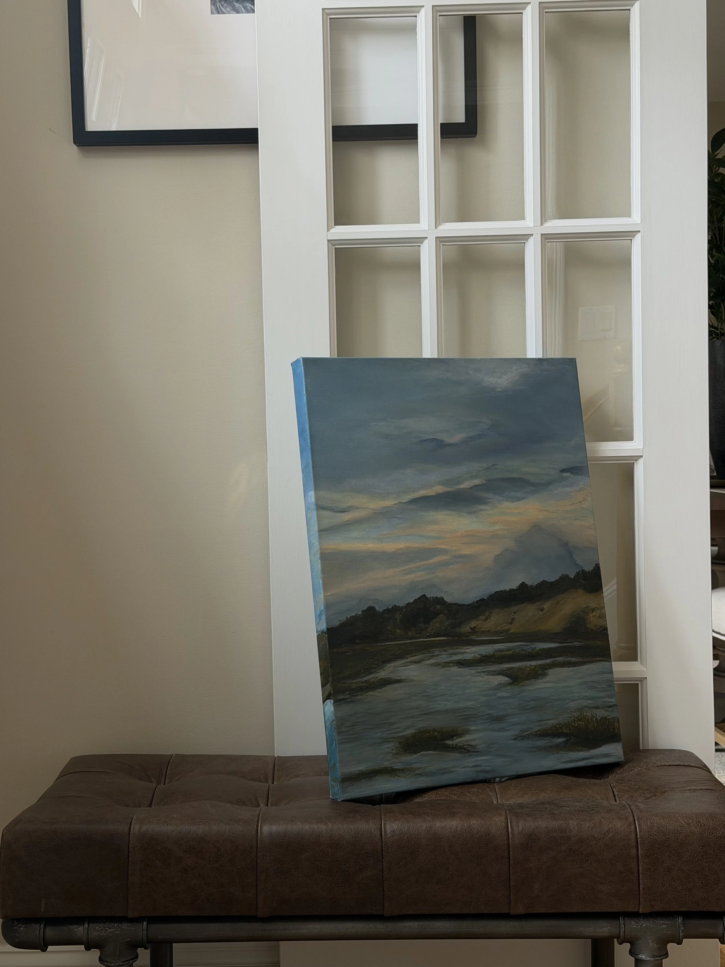 A painting of a landscape scene with mountains, sky, and water, leaning against a beige wall with a grid-shaped glass-paneled door in the background, placed on a padded brown bench.