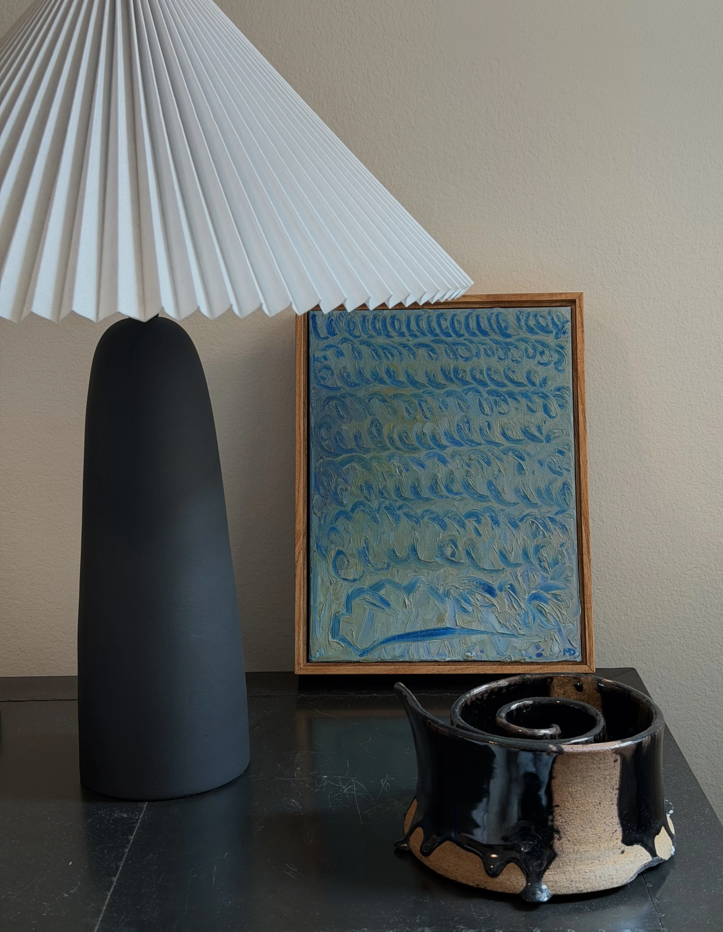 A beige and black ceramic cup with a wavy rim filled with a dark liquid, placed on a black surface in front of a framed abstract painting with blue and green swirls, and a tall black and white table lamp with a pleated white shade.