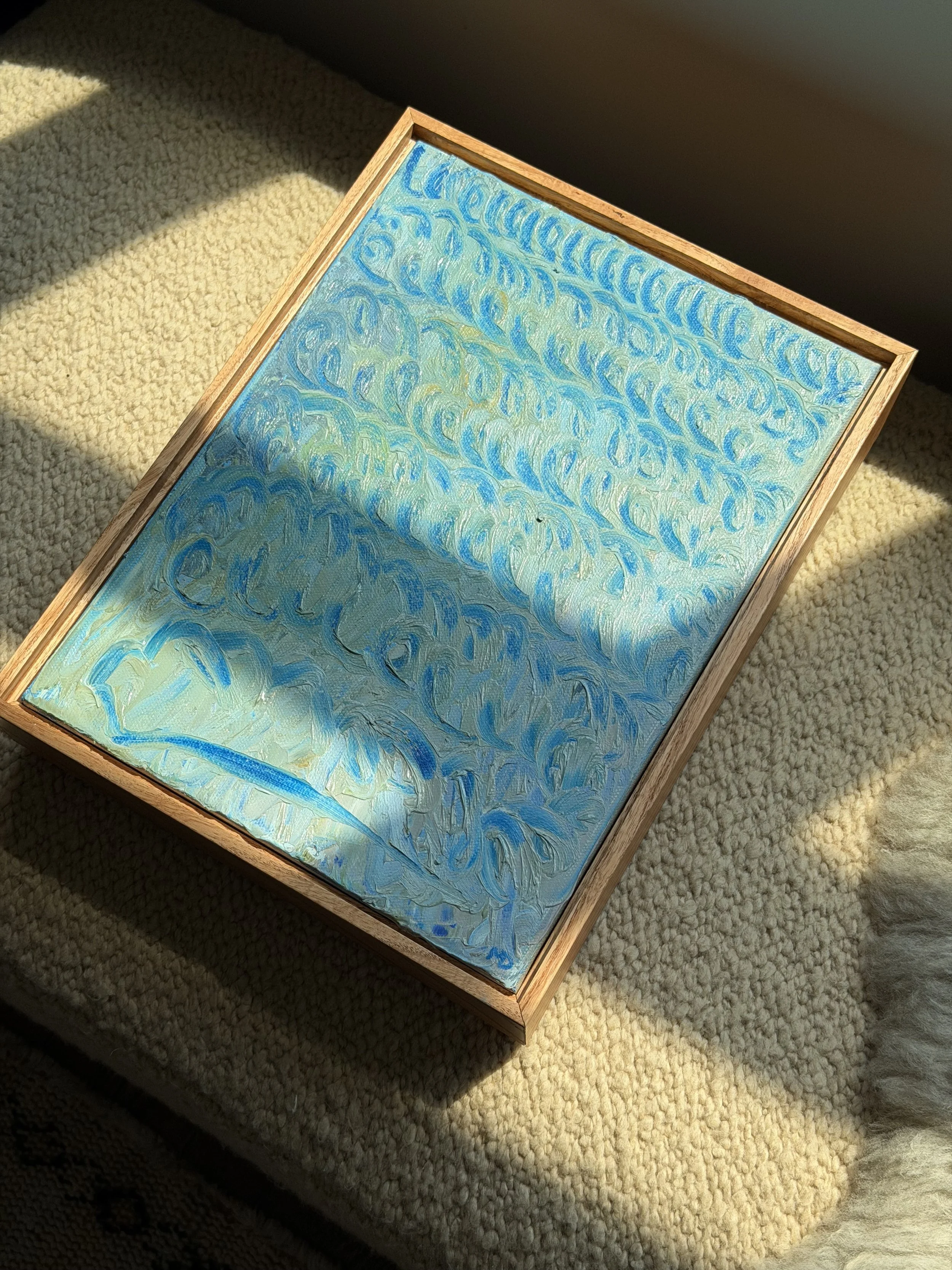 Close-up photo of a small painting with blue and green swirls, framed with a wooden border, placed on a beige carpeted surface in sunlight.