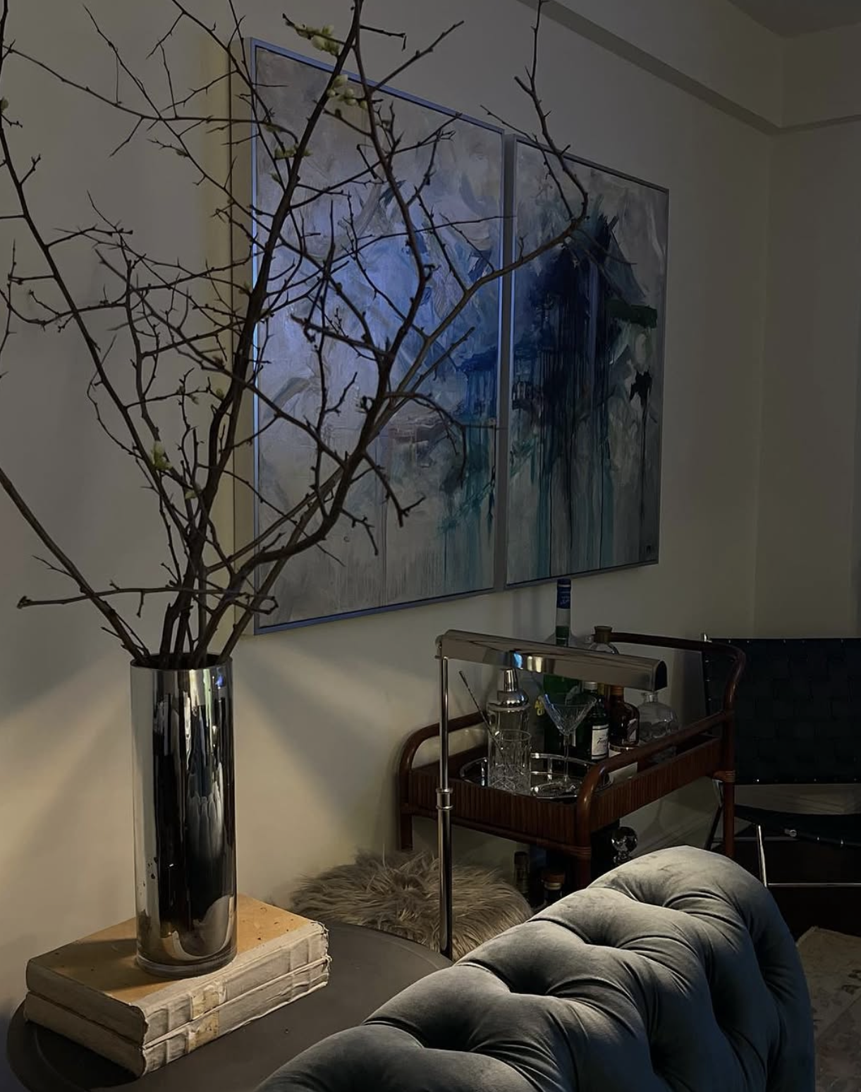 Interior of a room featuring a tall metallic vase with bare branches and small blossoms, hanging abstract artwork in shades of blue and gray, a bar cart with glassware and bottles, and a seating area with a tufted gray armchair and a black chair.