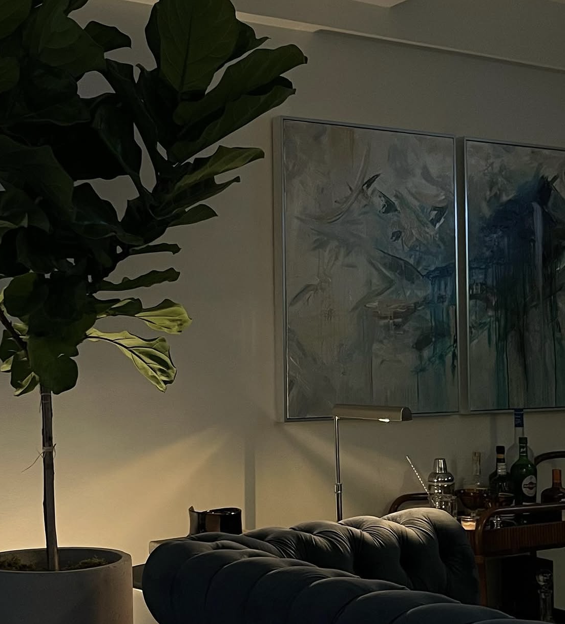 Interior of a living room with a large potted plant, a dark tufted sofa, a floor lamp, and a bar cart with bottles, glasses, and bar tools. There are two abstract paintings on the wall.