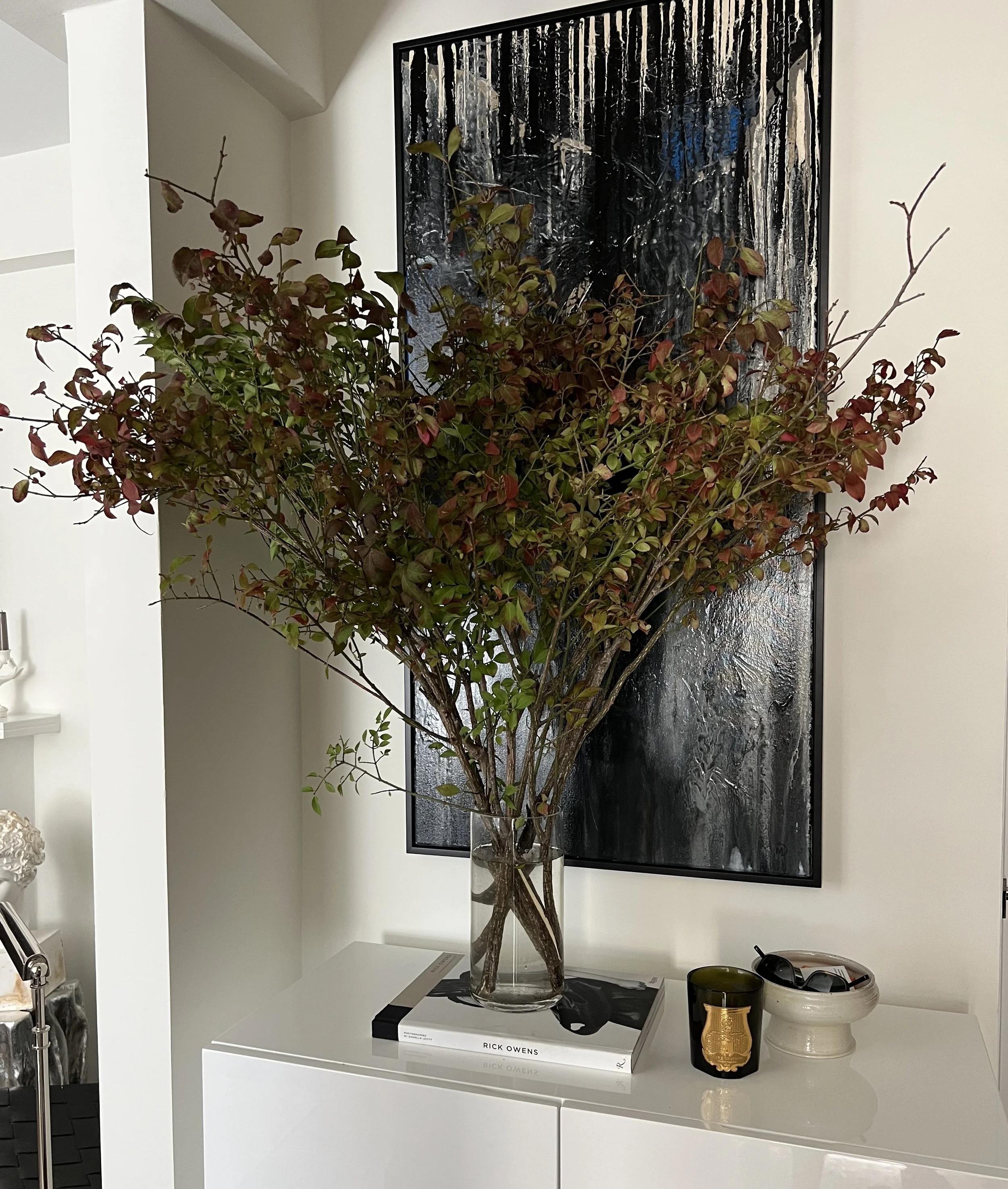 A large potted plant with reddish and green leaves inside a glass vase on a white surface, with a black and white landscape painting hung behind it. The plant is placed on a white table with a book titled "Rick Owens" beneath the vase, and there are 