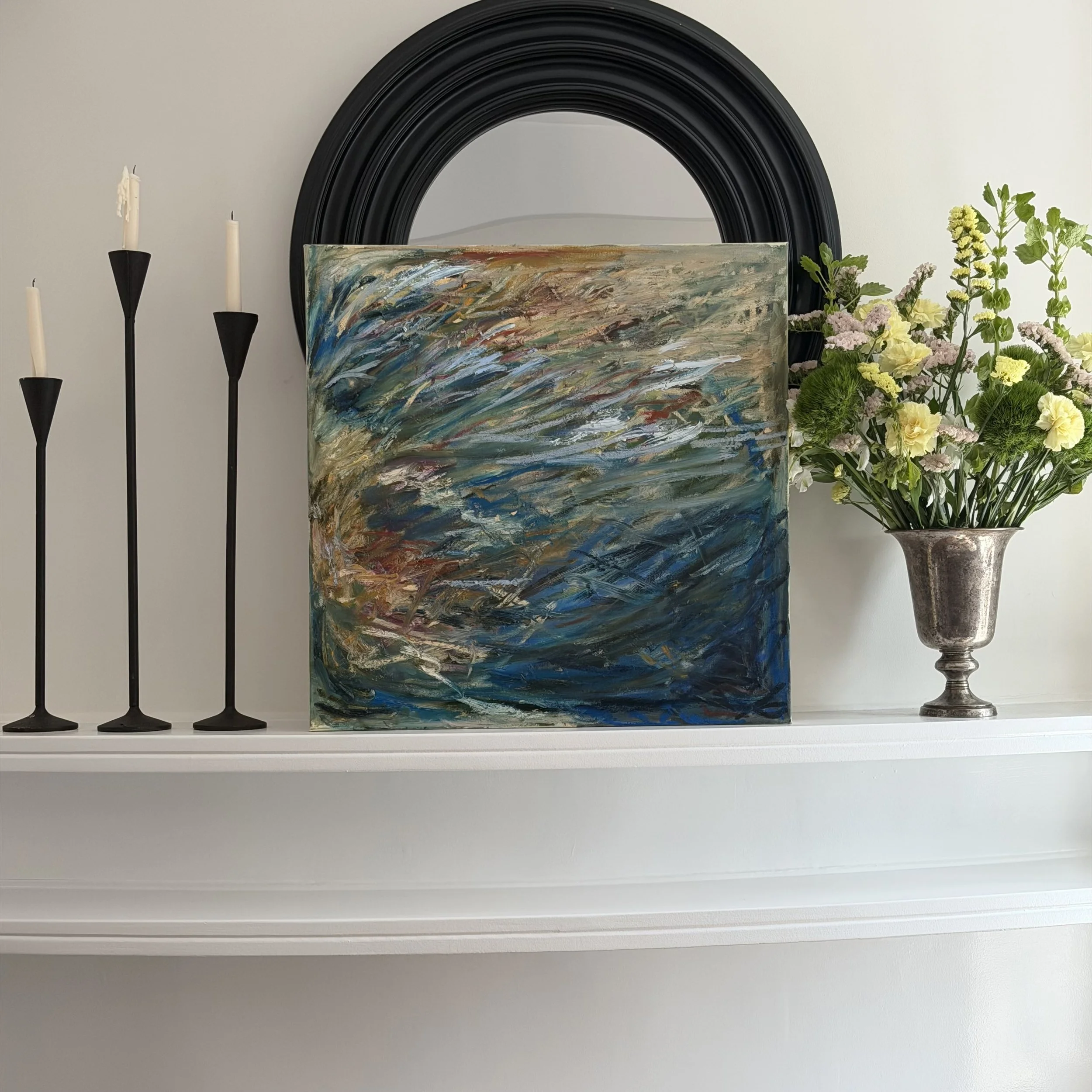 White shelf with a flower vase, several black candle holders with candles, an abstract painting, and a large black round mirror on the wall behind the objects.
