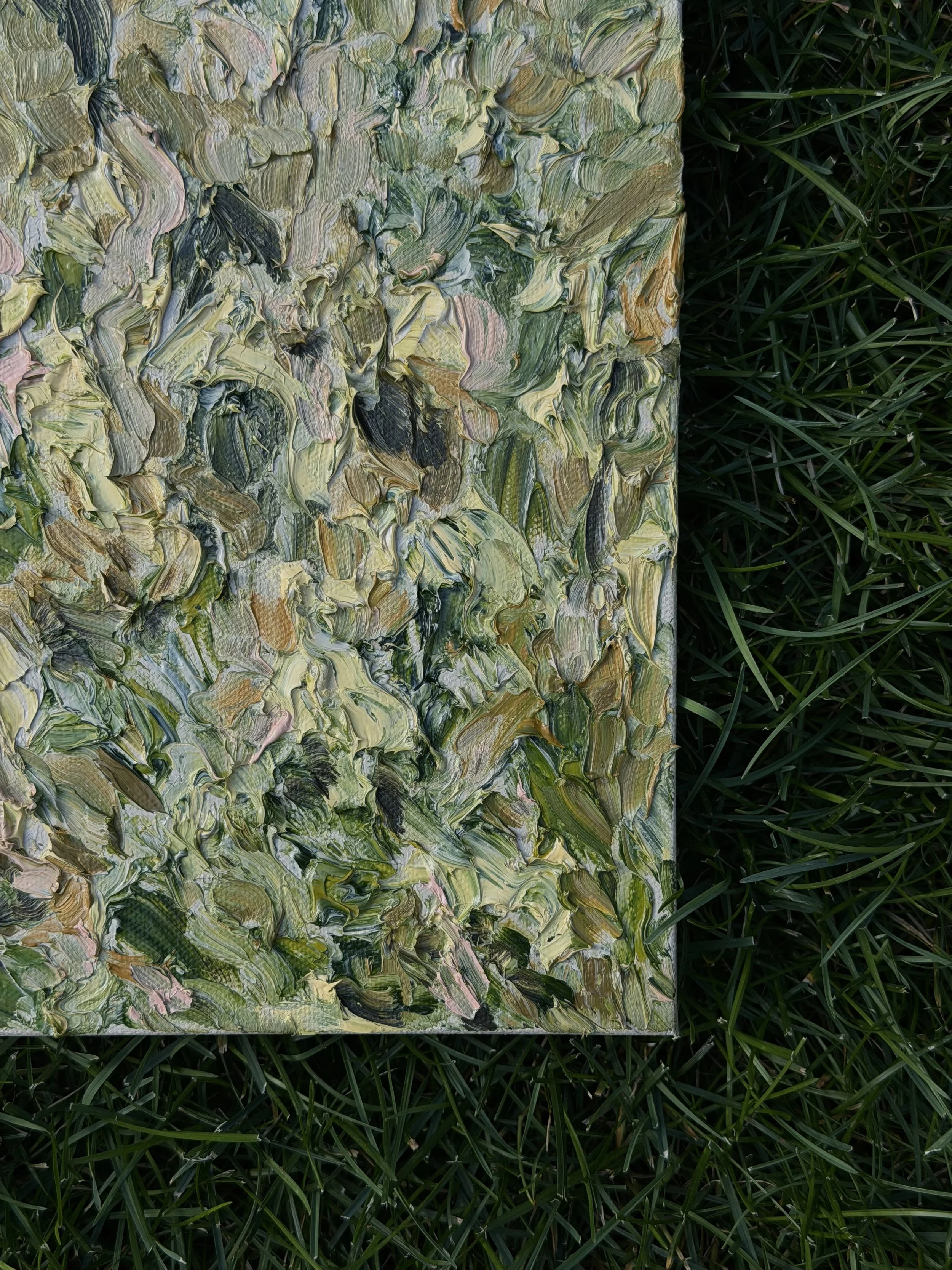 Close-up of an abstract oil painting with swirling brushstrokes in shades of green, beige, pink, and black, placed on grass.