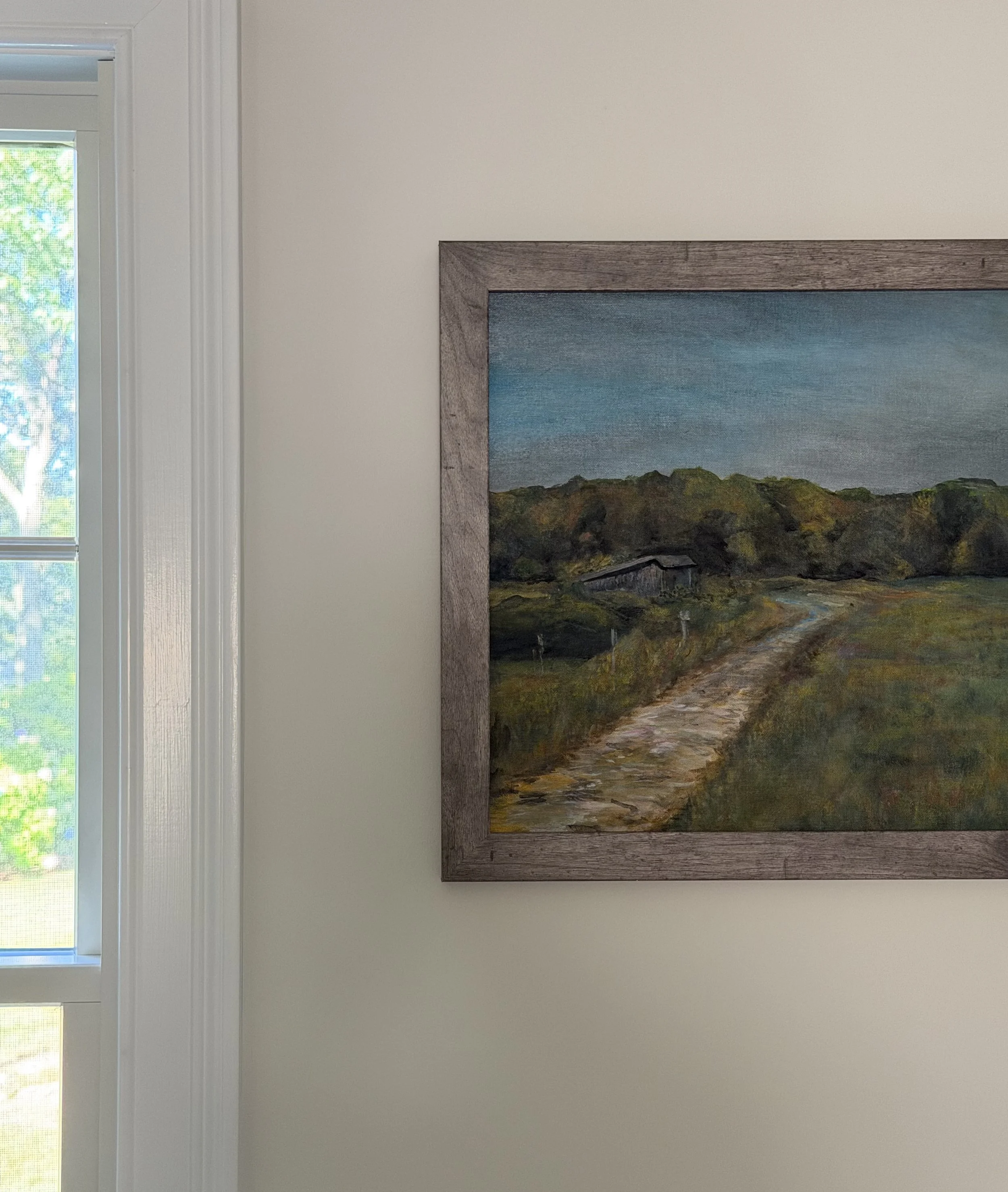 Interior wall with landscape painting in a wooden frame, and a window with blinds showing green trees outside.