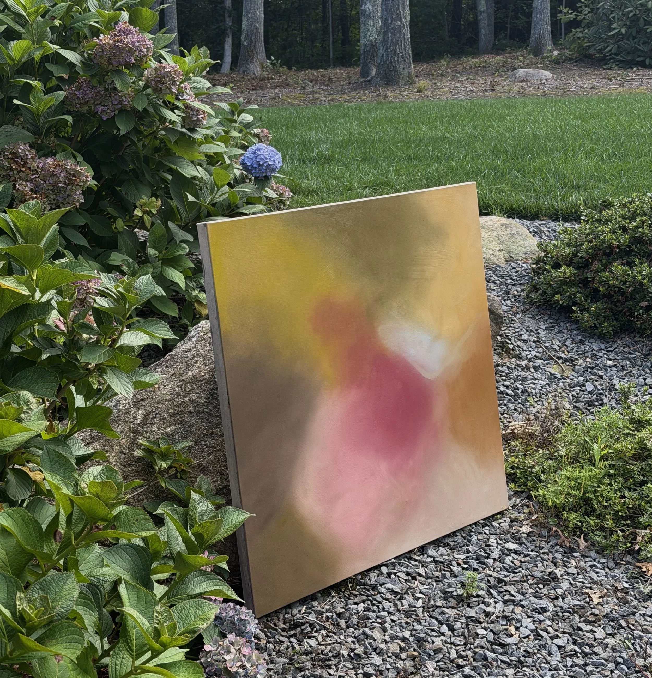 Colorful abstract painting on a standing canvas outside, surrounded by green plants, trees, and gravel.