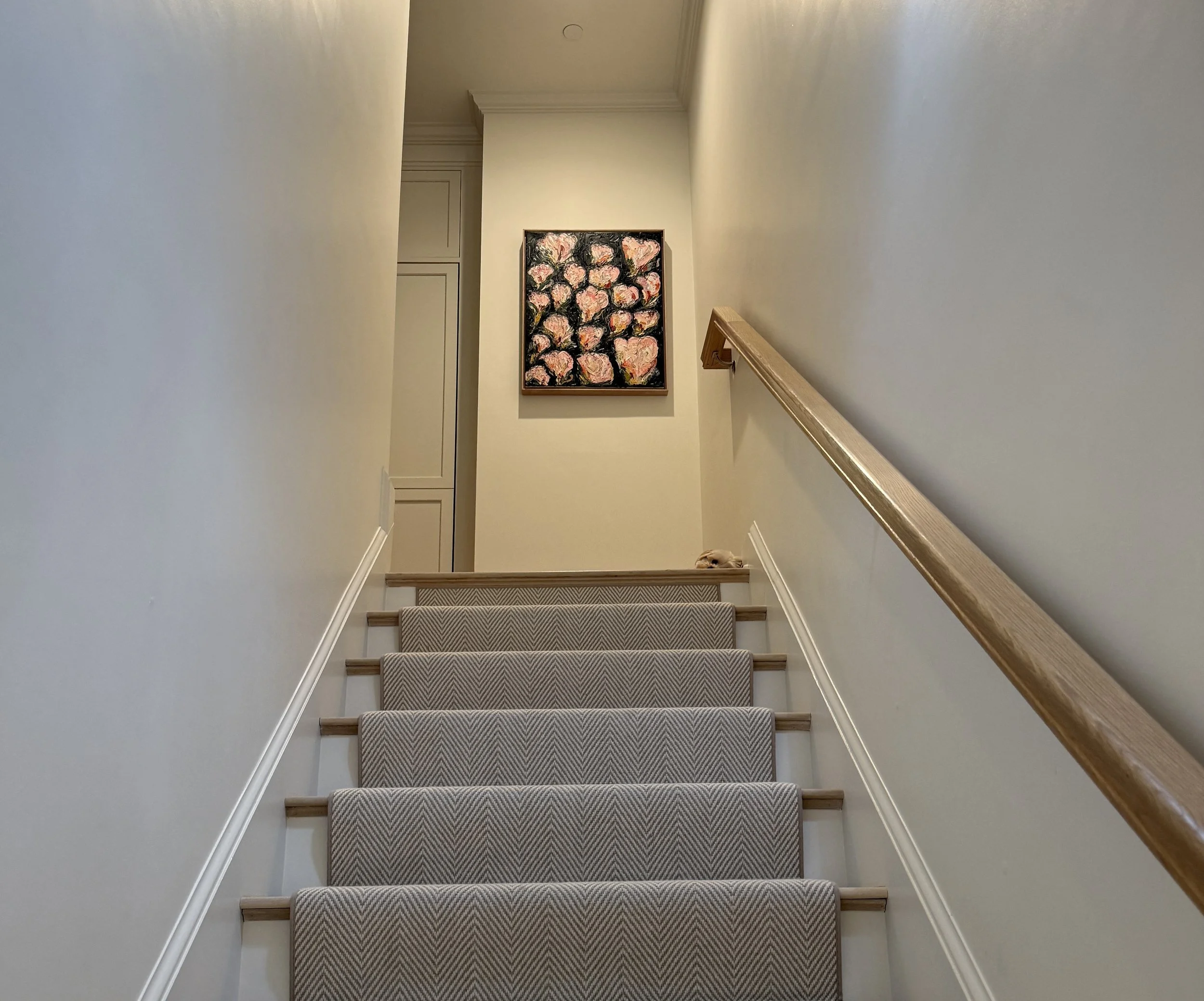 View of carpeted staircase with beige walls, wooden handrail on right, artwork with pink and white flowers on black background on the wall at the top of the stairs, and a dog peeking out from the bottom of the staircase.