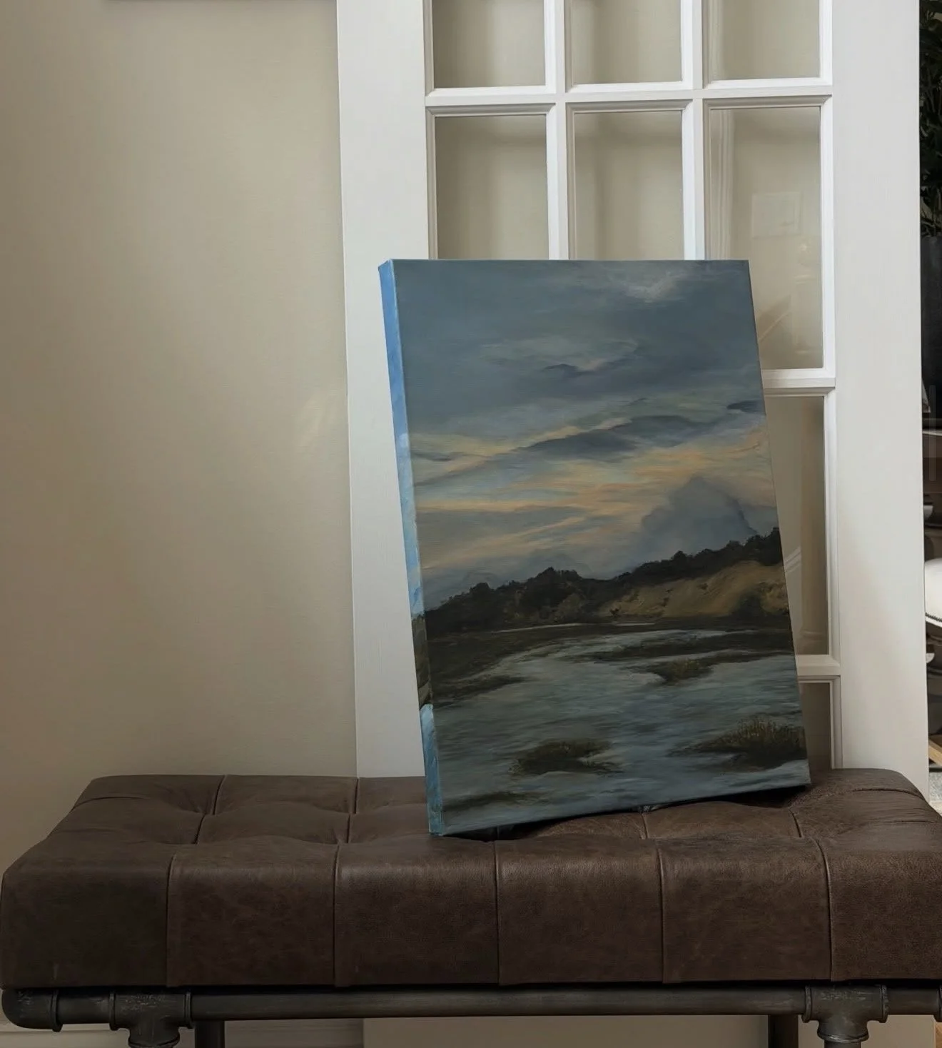 A landscape painting of a river, with a blue sky and clouds, leaning on a brown cushioned bench in front of a white decorative window frame.