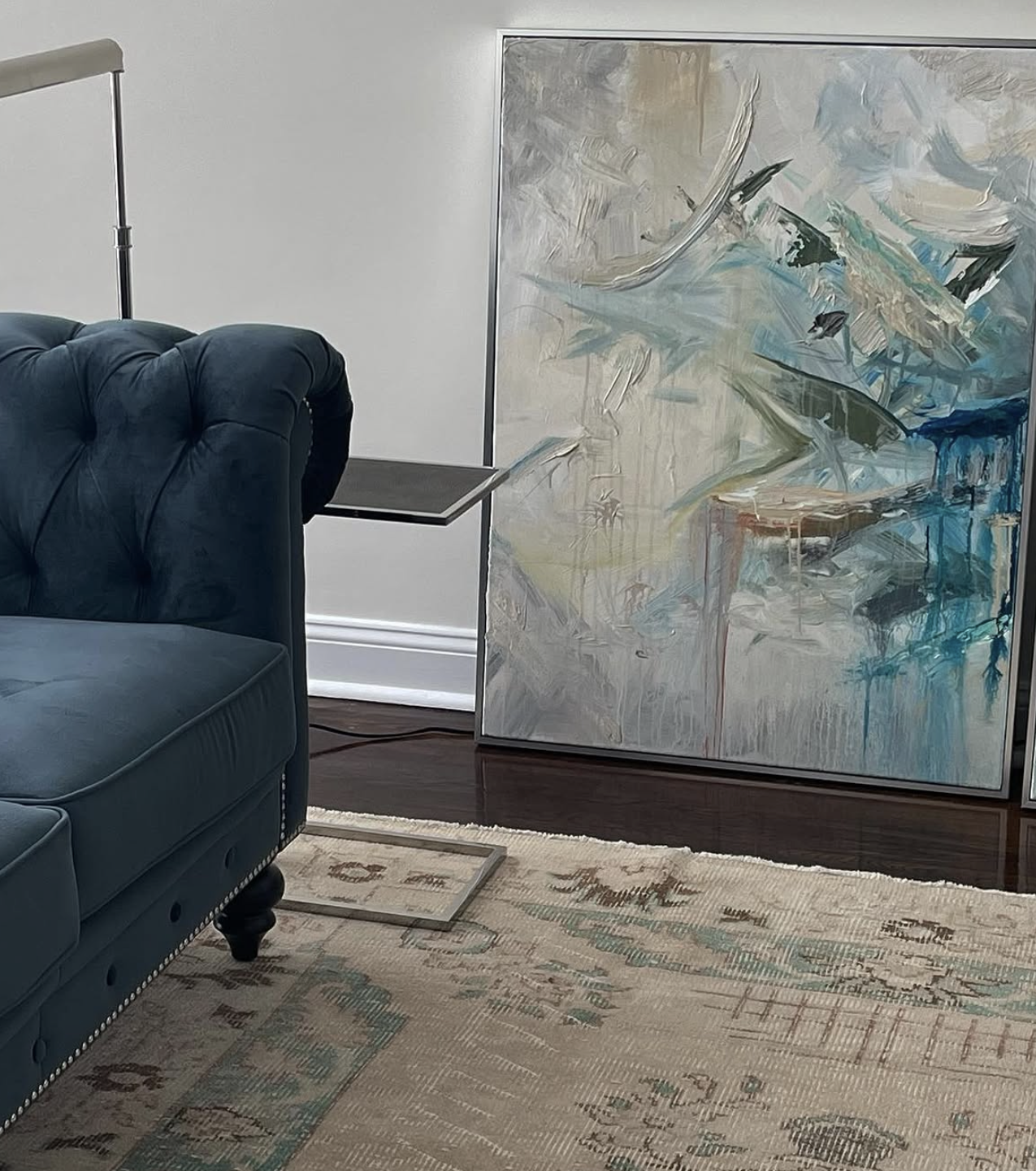 A modern living room corner with a navy blue tufted armchair, a beige patterned rug, a black side table, and an abstract painting with shades of blue, beige, and black leaning against the wall.