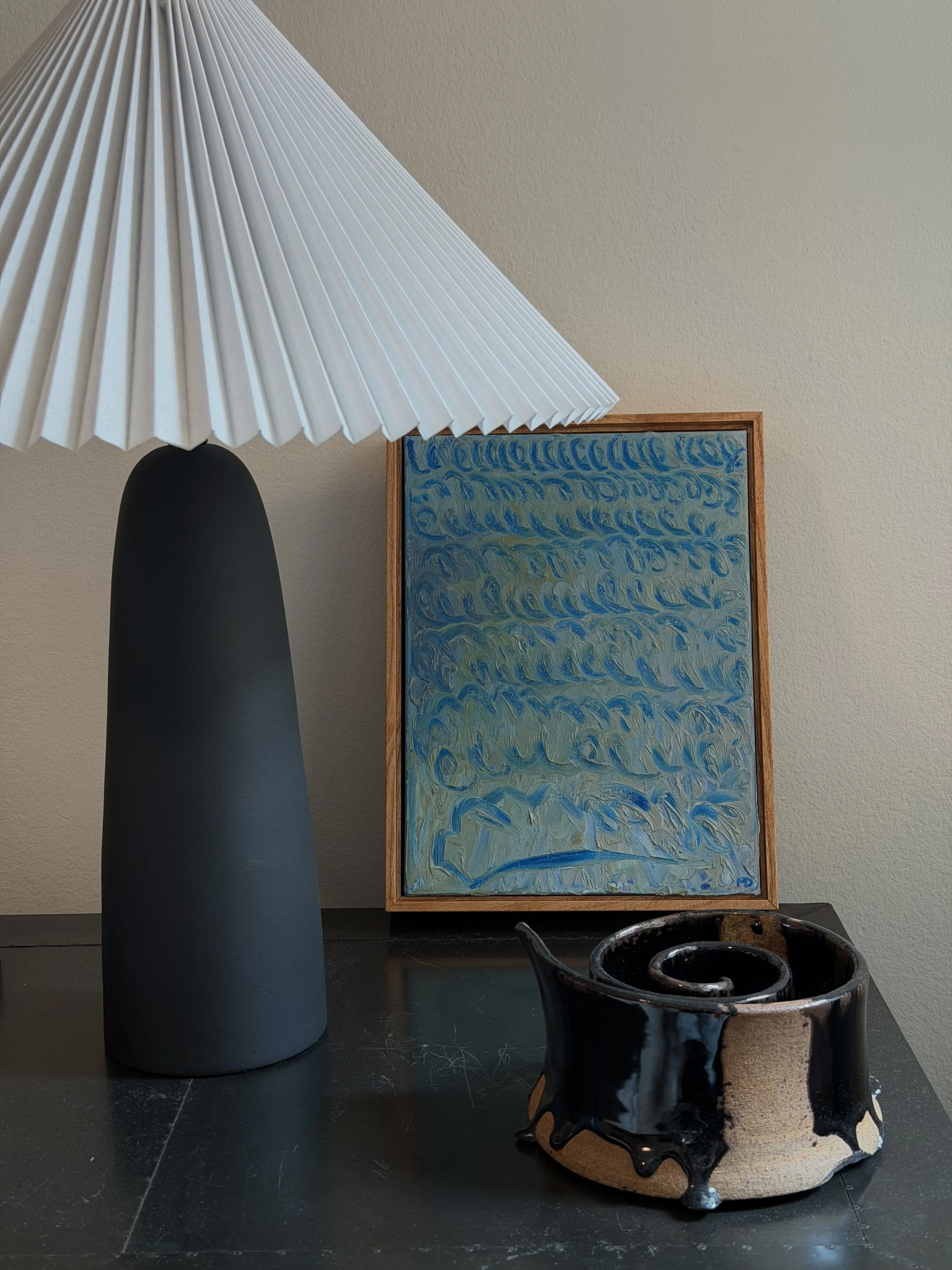 A table lamp with a black base and a large pleated white shade seen beside a framed abstract painting with blue and green wave-like patterns. In front of the painting, there is a black and beige ceramic pot with a glossy black glaze dripping down the