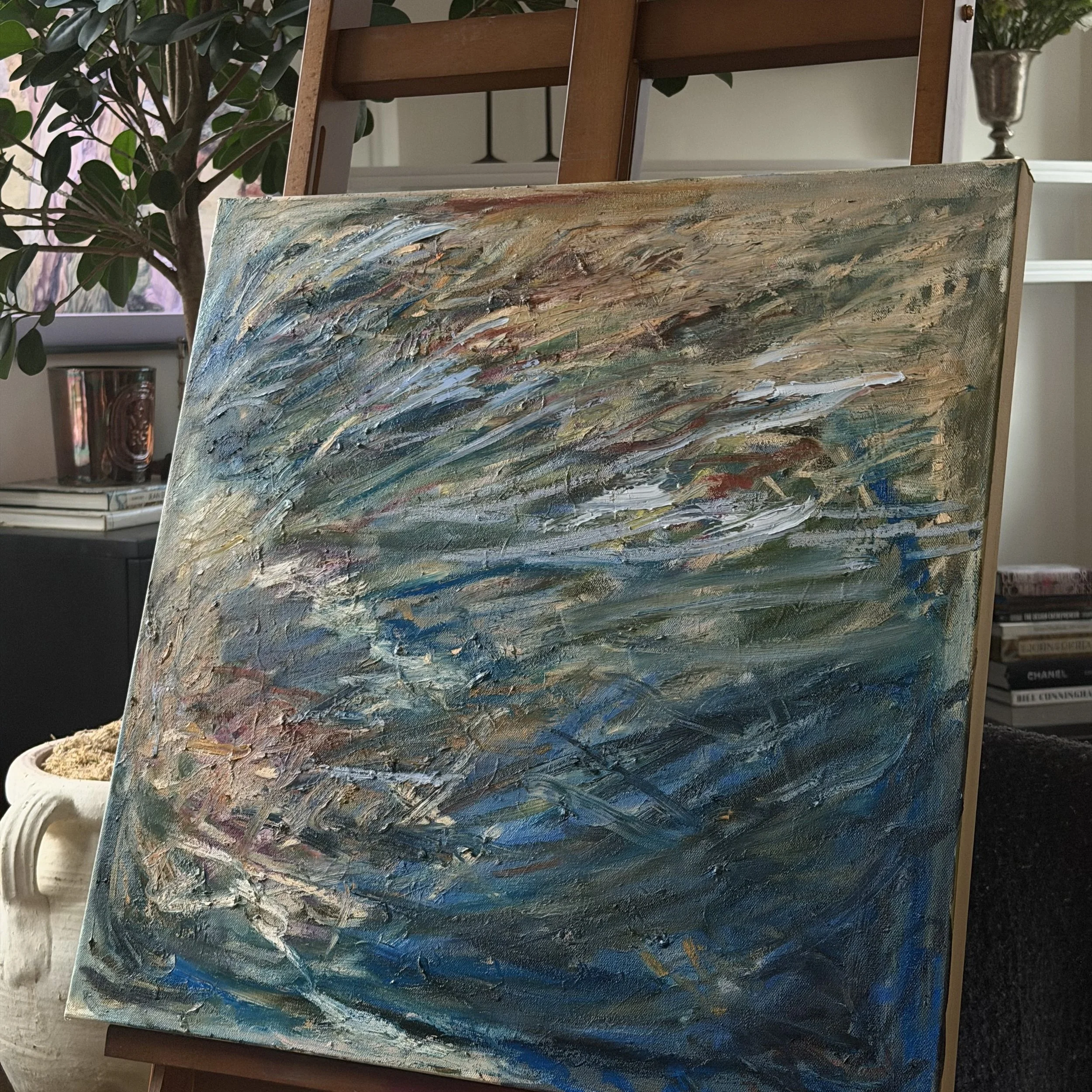 Close-up of an abstract oil painting on a canvas, featuring thick, textured brushstrokes in shades of blue, green, beige, and hints of red and white.