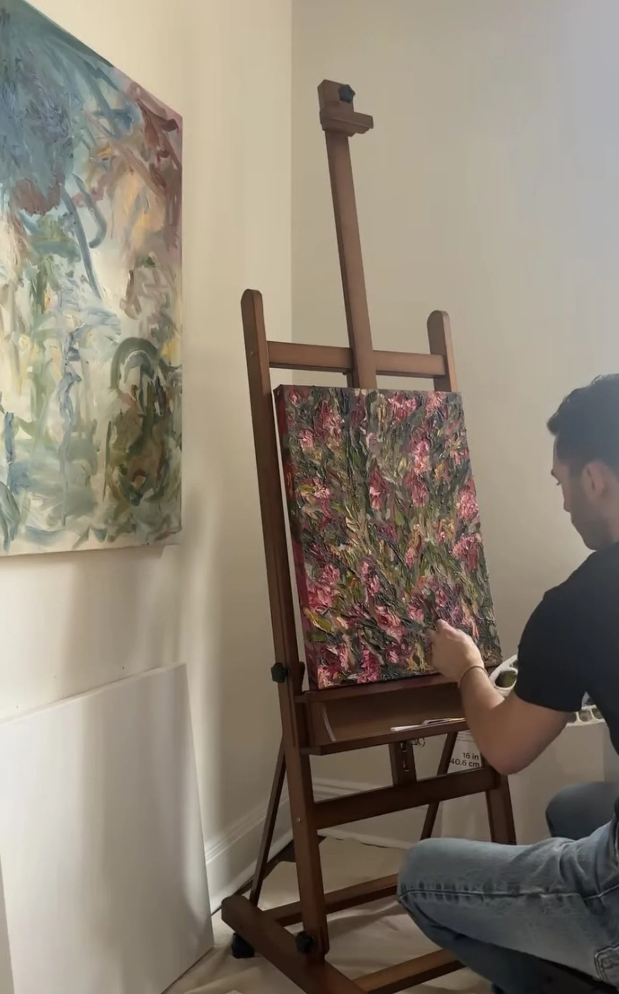 A person painting an abstract floral artwork on a canvas set on an easel in a room with white walls and wooden flooring. Another large abstract painted piece hangs on the wall to the left.