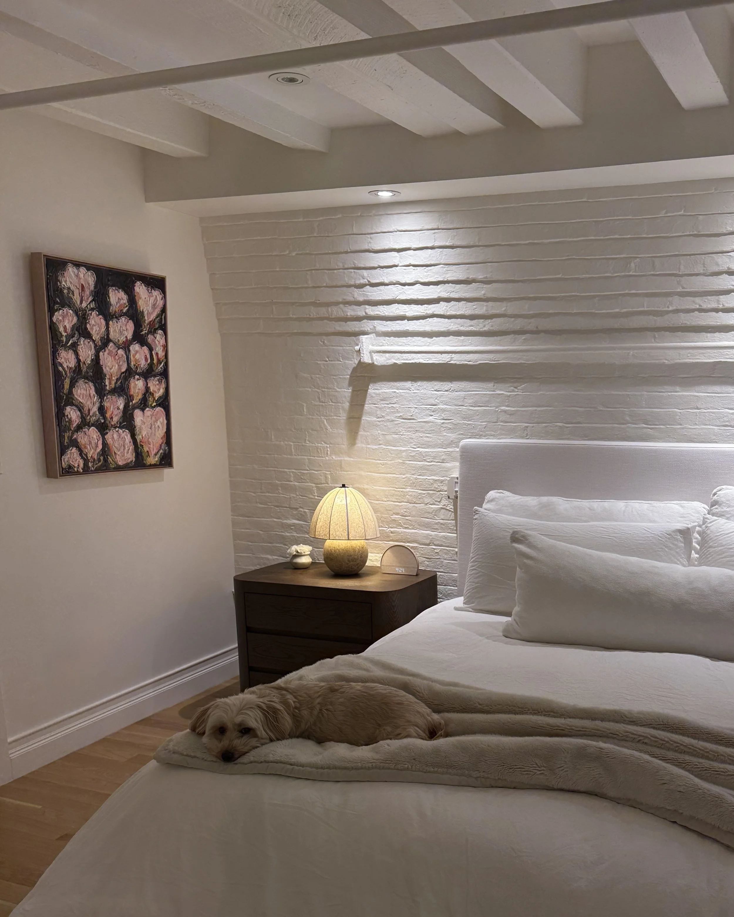 A cozy bedroom with white brick walls and wooden flooring. There are two beds with white bedding, and a bedside table with a lamp. A dog is lying on a cushion at the foot of one of the beds. There is a painting of pink roses on the wall.