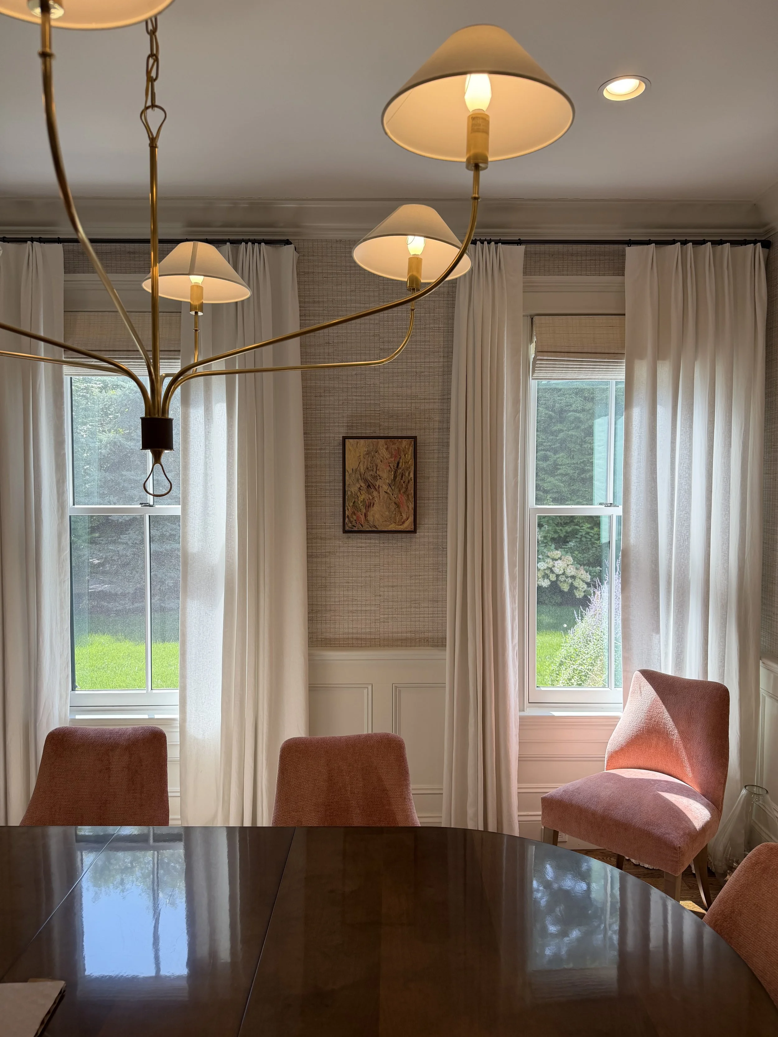 Dining room with a round dark wood table, four pinkish chairs, large windows with white curtains, a framed painting on the wall, and a chandelier with multiple lampshades hanging from the ceiling.