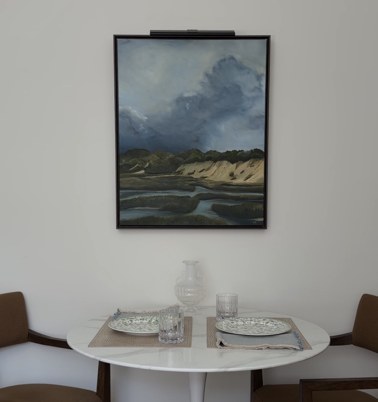 Interior view of a dining area with a white oval table set with placemats, glasses, and plates. There are two brown chairs on each side. On the wall behind the table, there is a large landscape painting of a cloudy sky over a grassy landscape with a 