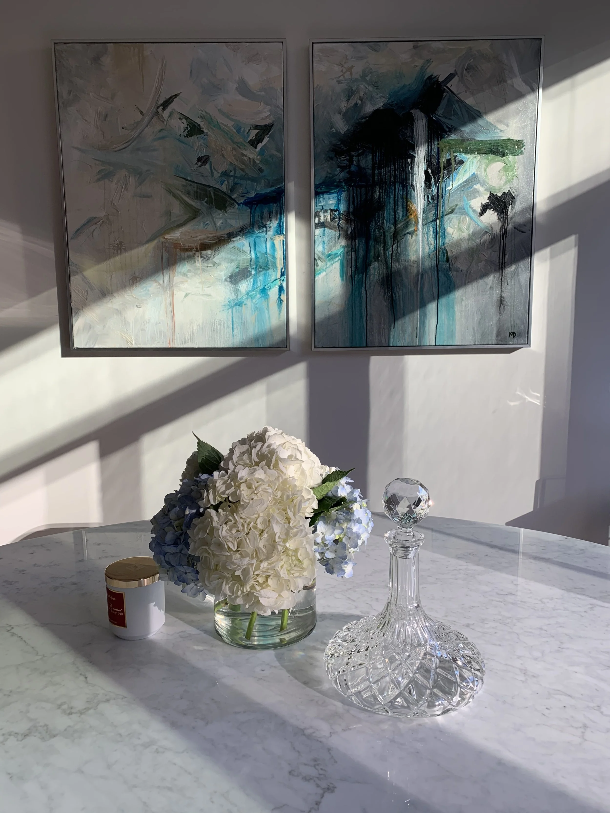 A marble table decorated with a white hydrangea flower arrangement in a glass vase, a crystal decanter, and a jewelry box. Two abstract paintings hang on the wall behind, with sunlight casting shadows across the scene.