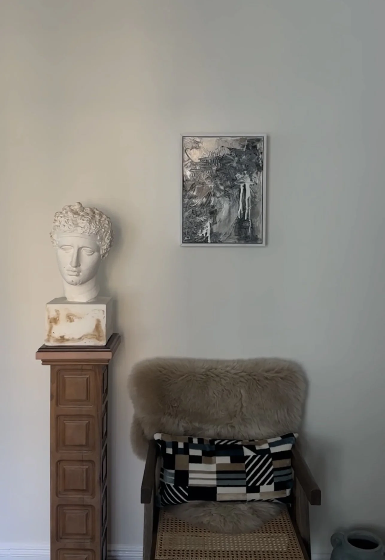 A white classical bust sculpture of a head on a tall wooden cabinet, a geometric-patterned chair with a fur cushion, a framed abstract black and white painting on a plain light-colored wall.