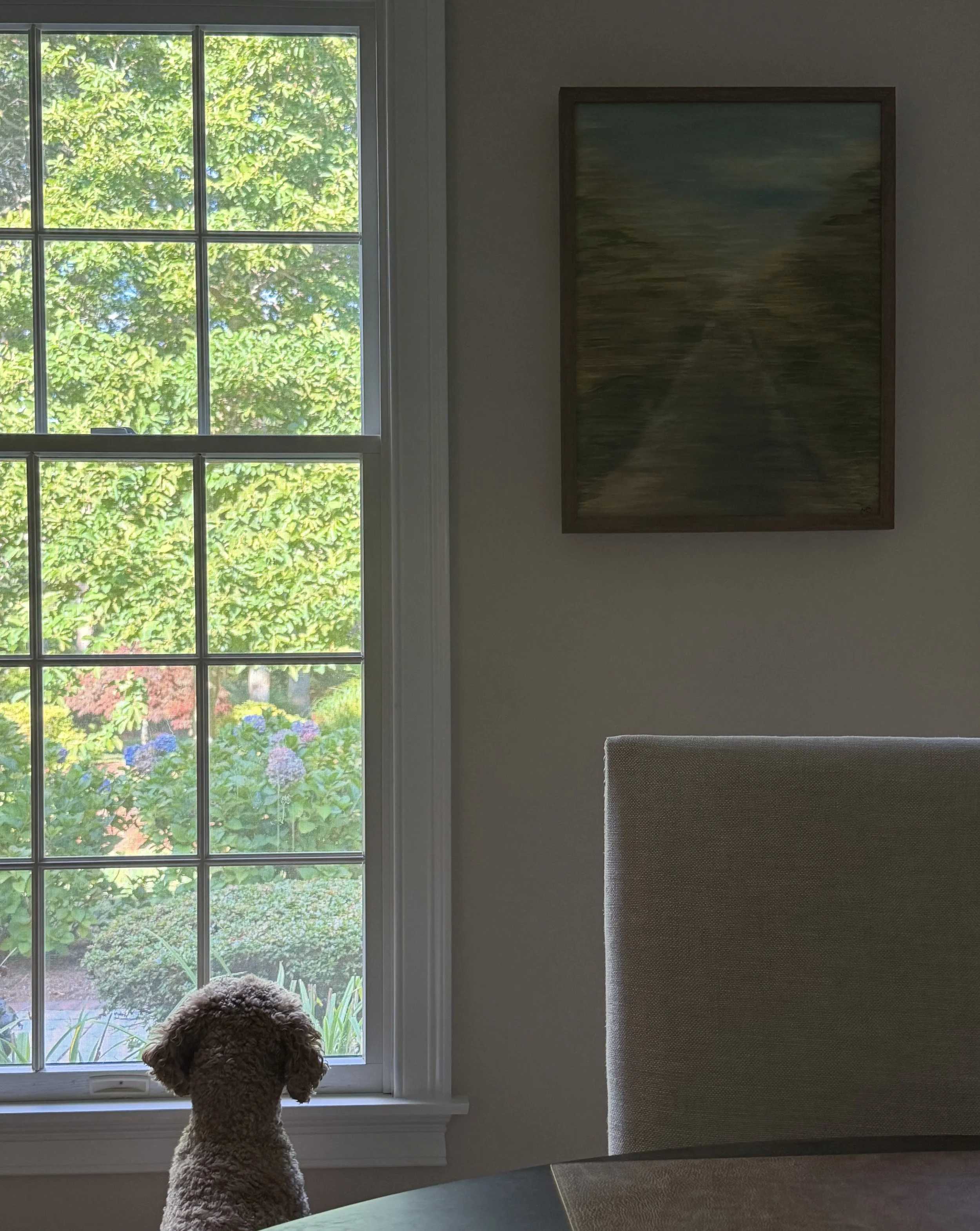 A brown curly-haired dog looking out a window at a lush green garden with trees and flowers.