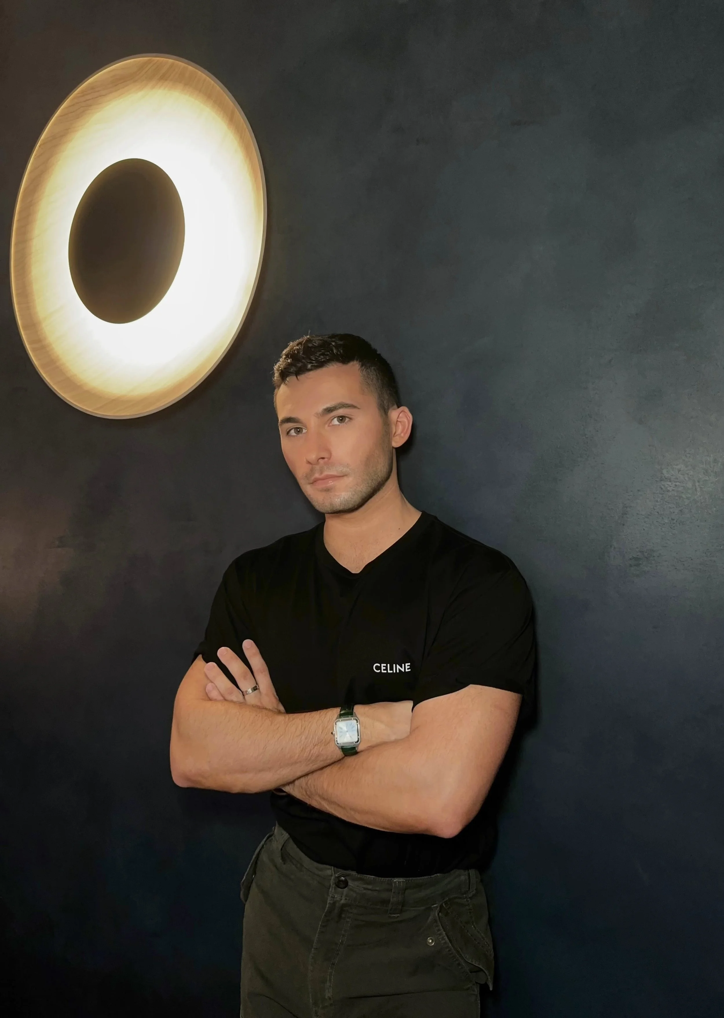 A young man with dark hair and light skin, wearing a black t-shirt with 'CELINE' printed on it, standing against a dark wall with a large circular light fixture overhead, arms crossed and looking at the camera.