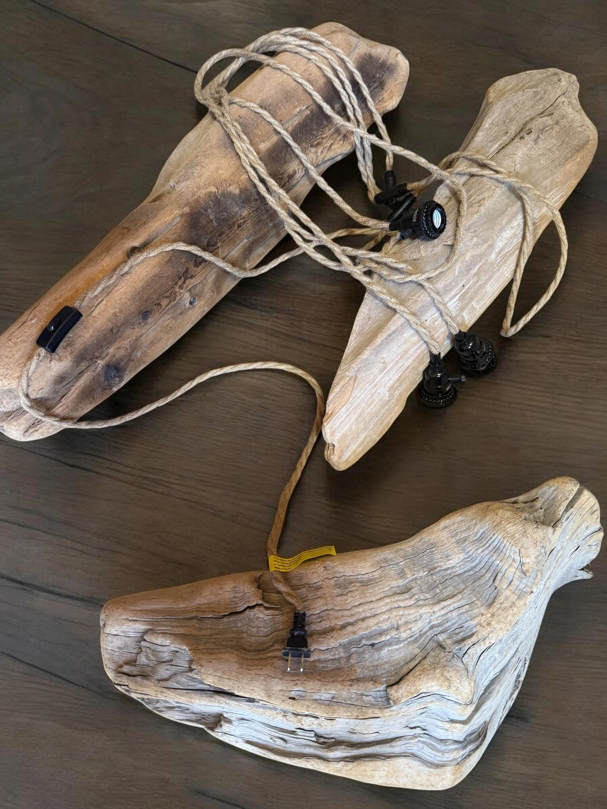 A long, twisted wooden piece with a light natural finish, placed on a dark wooden surface, with black and yellow electrical cords and black connectors draped over it.