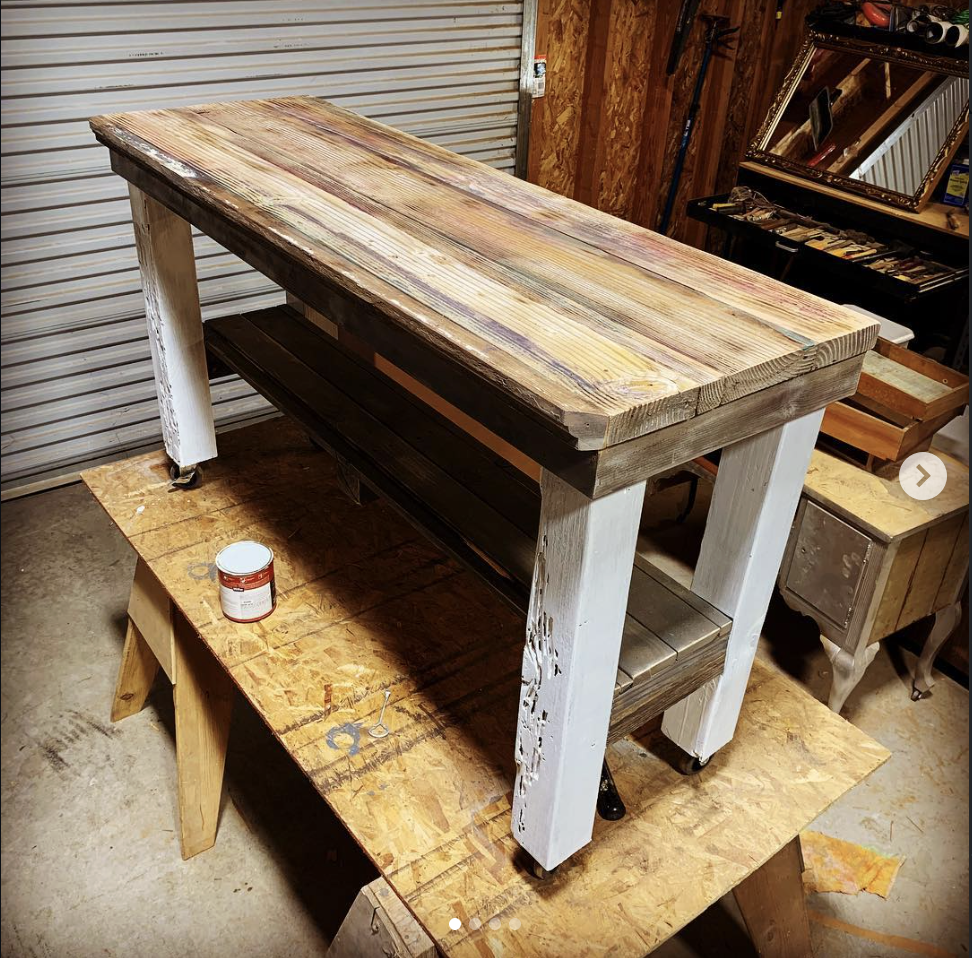 A wooden workbench with a rustic, wooden top and white painted legs, situated in a woodworking or garage workshop for sale.