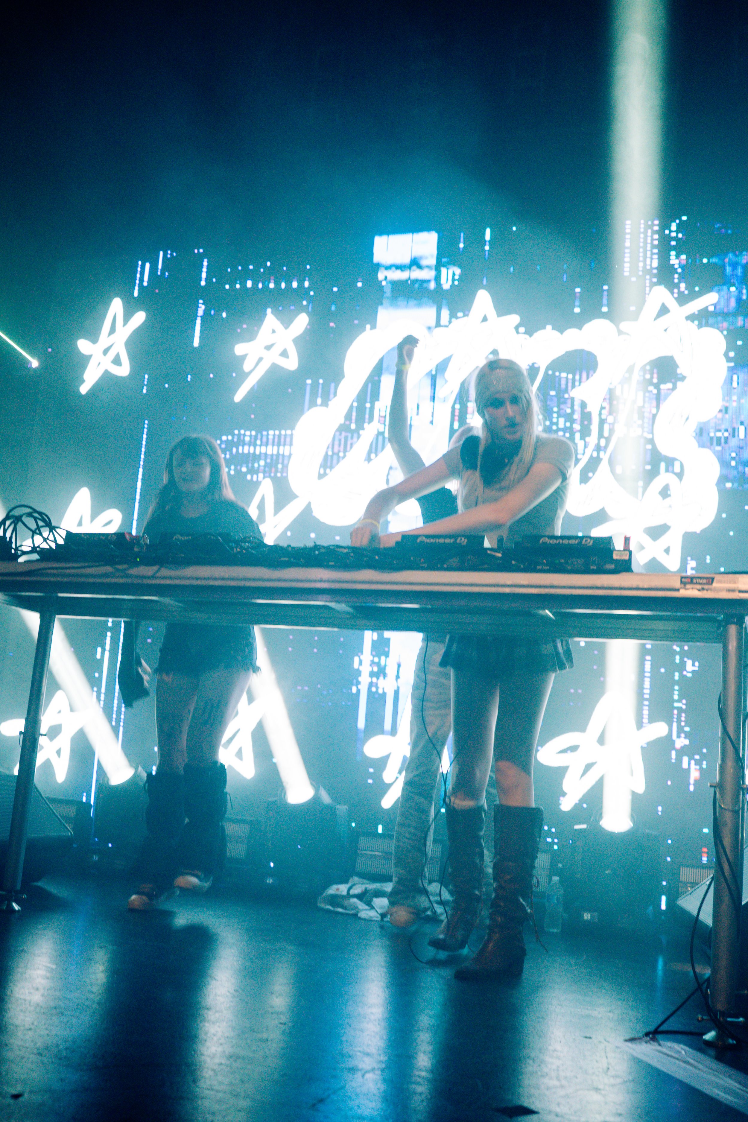 Two female DJs perform on stage with a large LED screen behind them displaying white handwritten text and illustrations, illuminated by vibrant blue stage lighting.