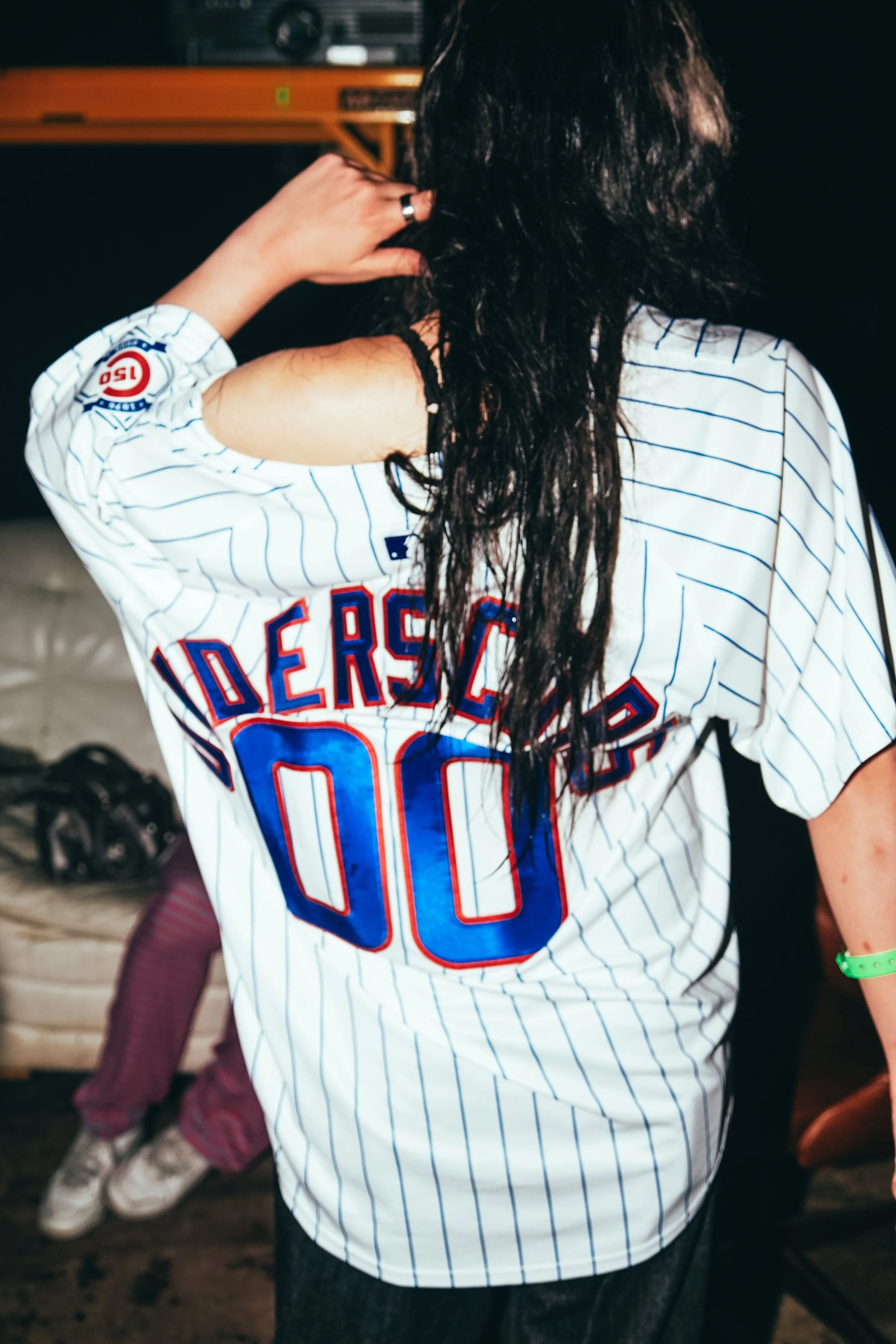 Back of a person wearing a white baseball jersey with blue lettering and the number 00. The person has long dark hair and is touching their head with one hand.