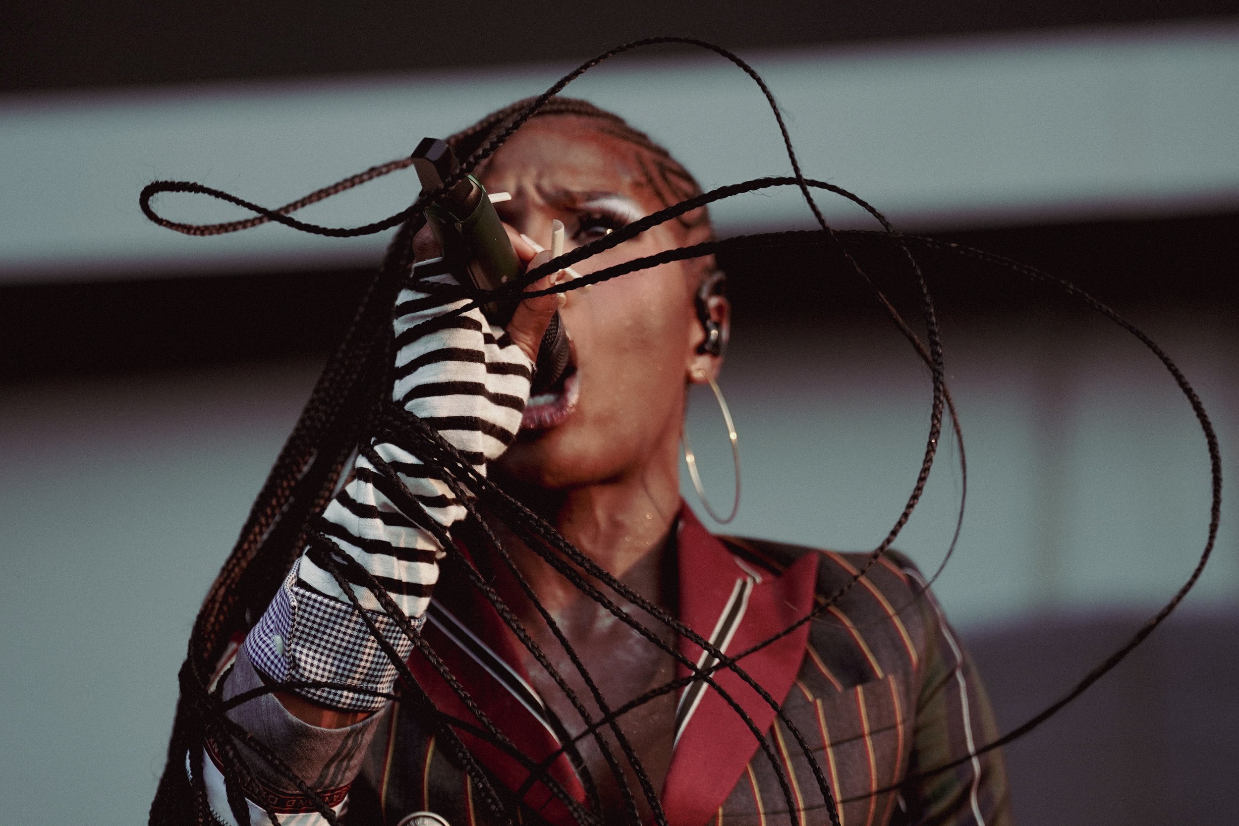 A woman with braided hair, talking on a mobile phone, behind a black wire fence. She is wearing a striped shirt and headphones, with earrings, and has a serious expression.