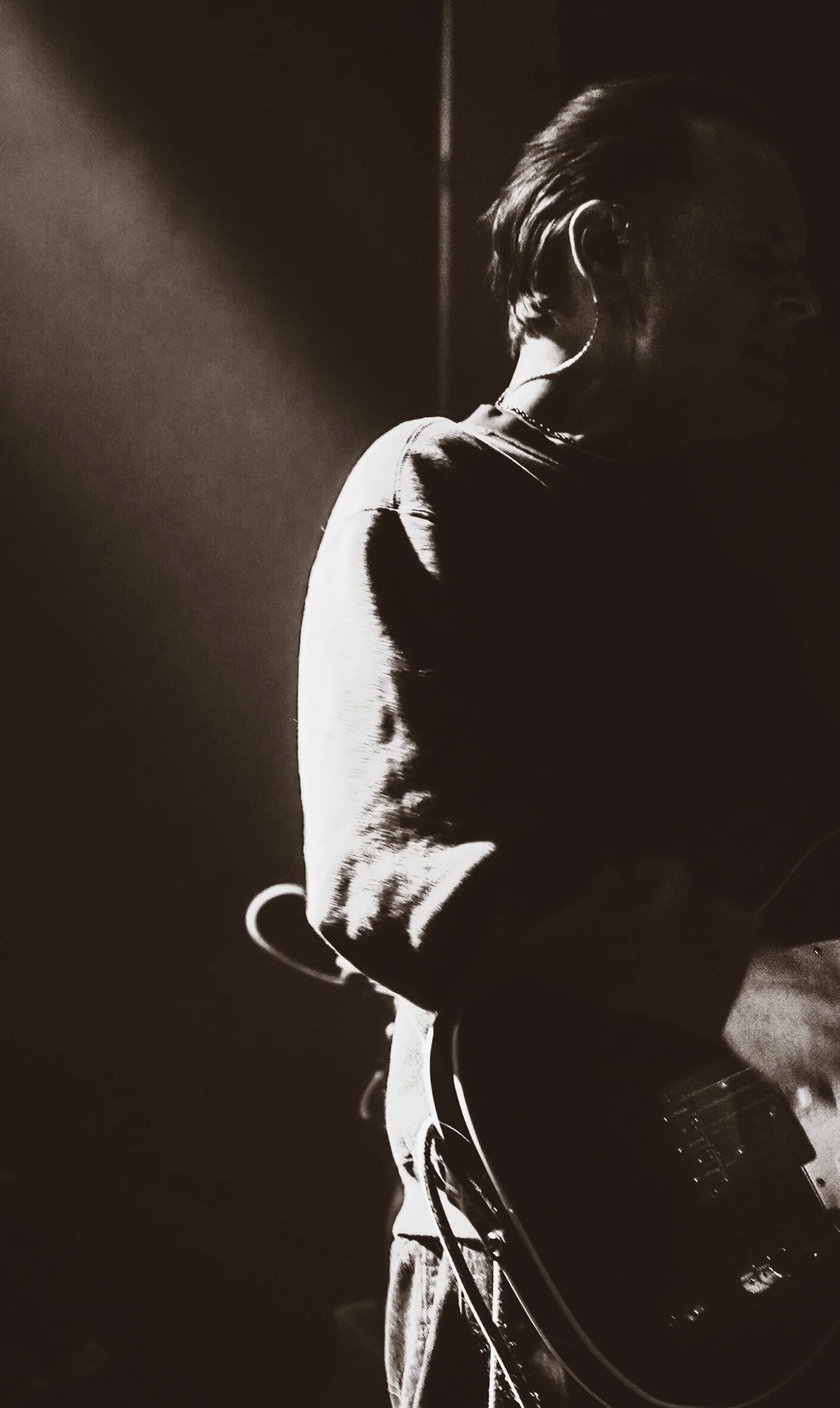 Silhouette of a person with short hair, facing sideways, holding a guitar in a dark setting with a light source highlighting parts of their face and clothing.