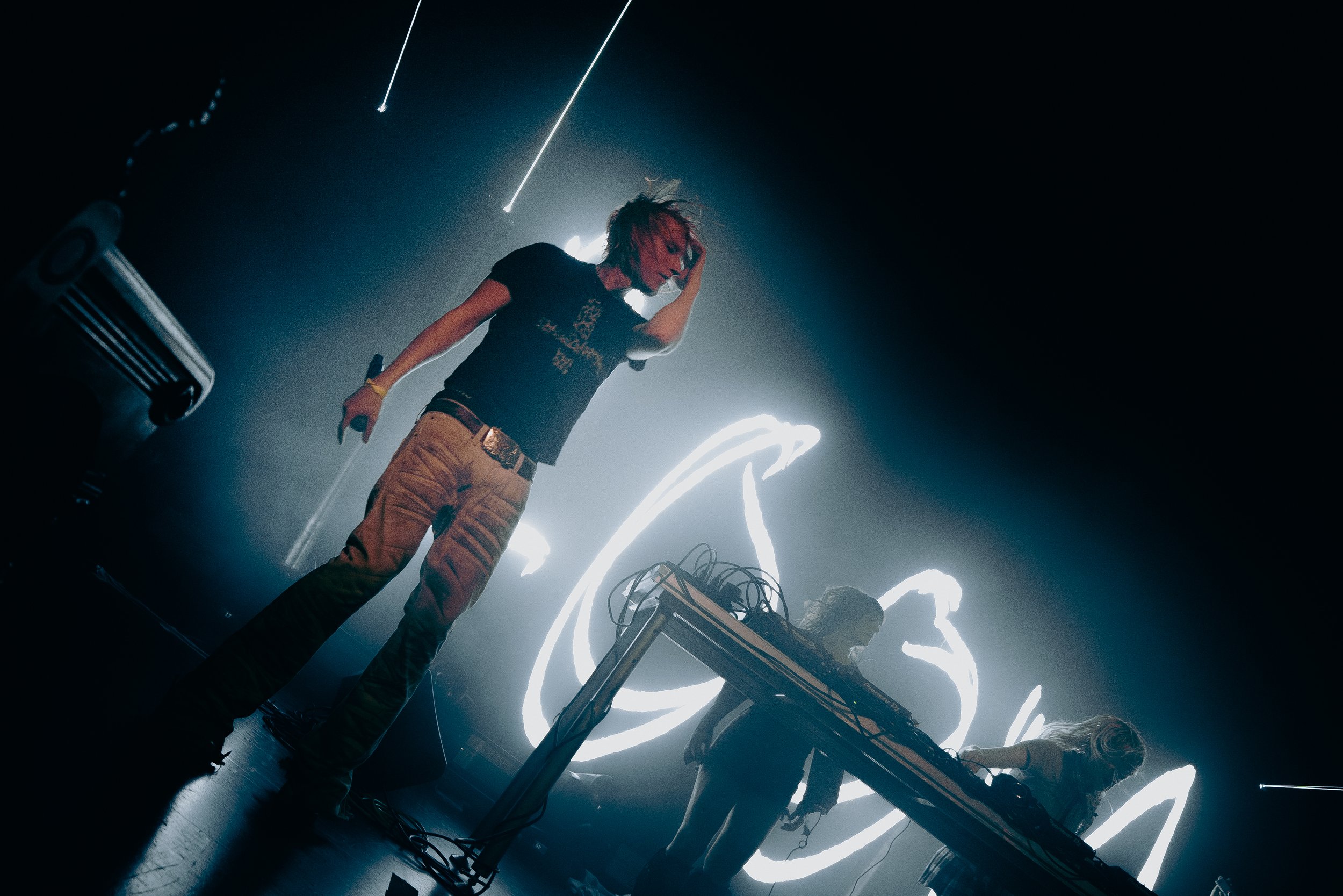 A performer with a face painted in red and black, wearing a black t-shirt and yellow pants, holding a microphone, standing on a stage with flowing light trails behind them, and a person in the background working with electronic equipment.