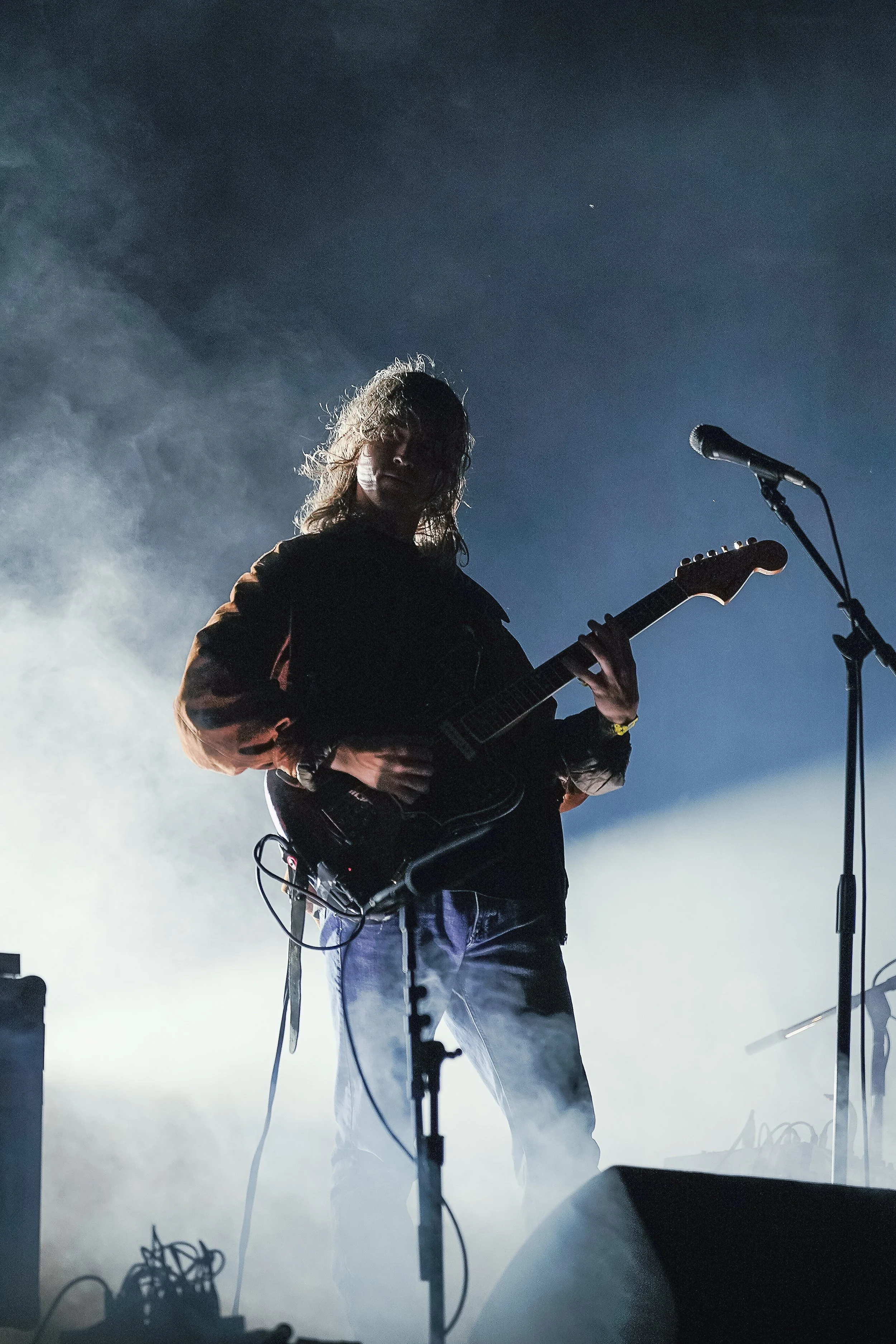 A person playing an electric guitar on stage with fog and a microphone nearby, backlit and partially shadowed.