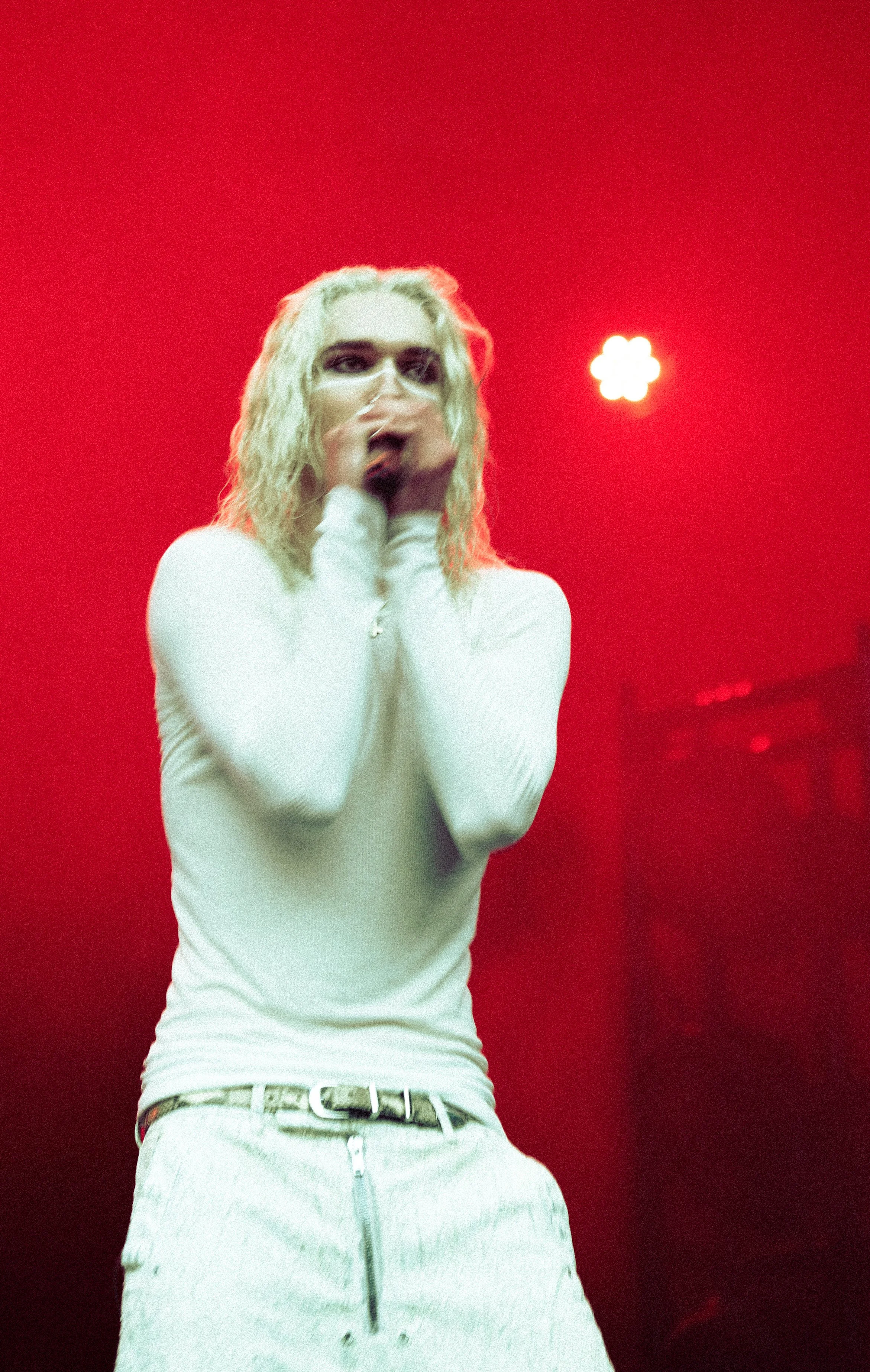 A person with long, bleached hair and dark makeup on their eyes, wearing a white long-sleeve shirt and light-colored pants with a belt, holding a microphone against a red background with a bright light