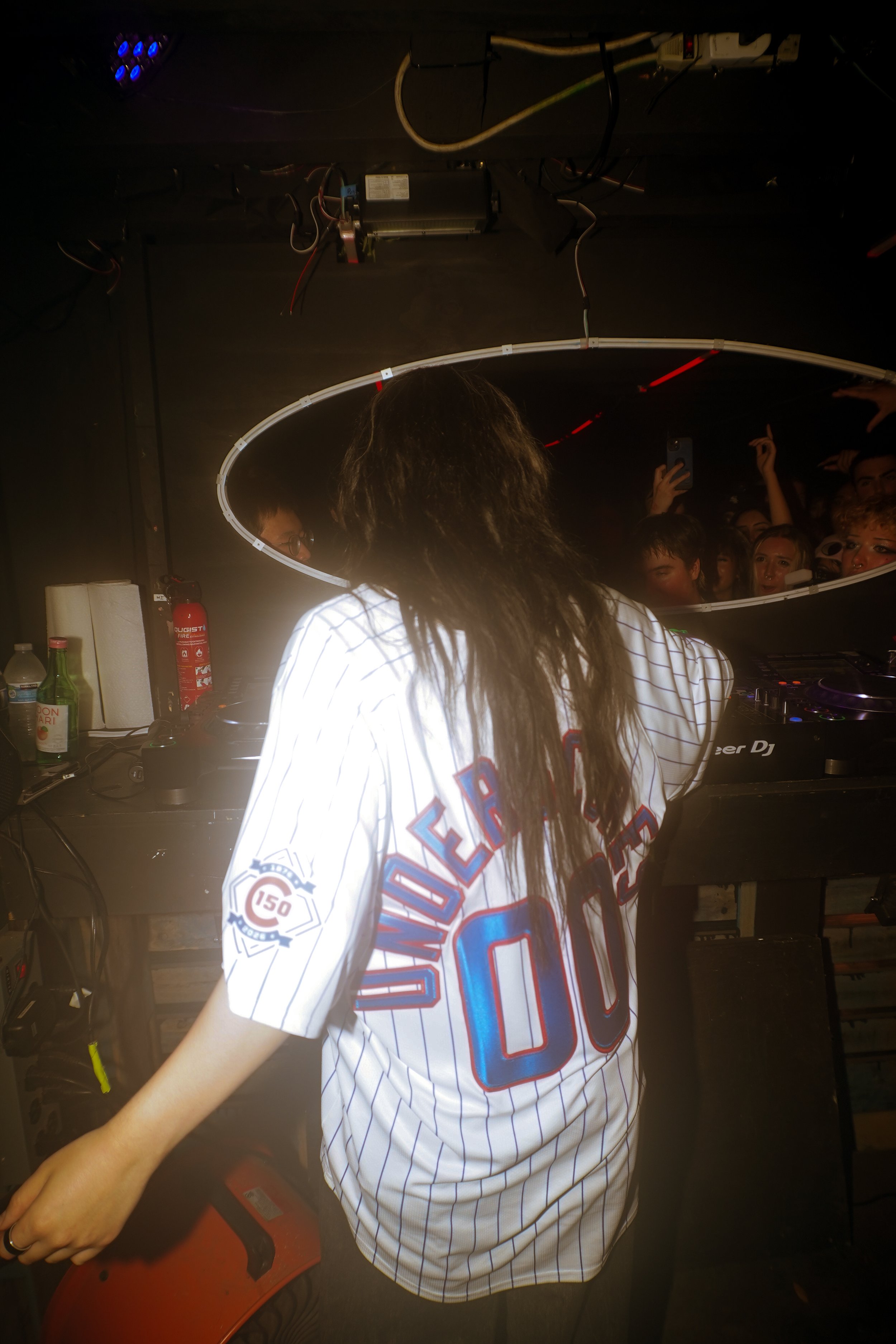 A DJ with long hair visible from behind, wearing a white sports jersey with blue and red accents, playing music at a dark venue. The crowd is visible in a mirror reflection above the DJ's head, with people raising hands and taking photos. DJ equipmen